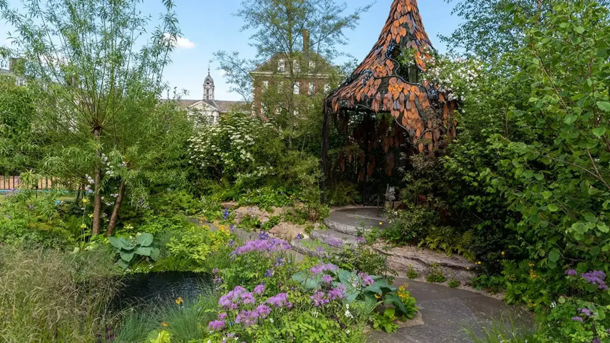 A sanctuary garden featuring a sculptural, leaf-patterned metal pavilion, a dark pond, and a winding stone path surrounded by lush green foliage and purple flowers.