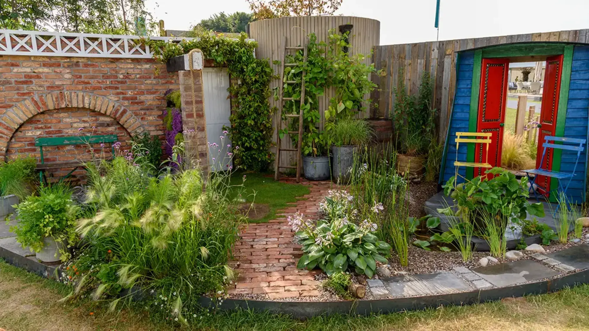 An eclectic garden with a vibrant shed, a timber structure, a brick wall and bench, a brick path, lush planting, and a small pond.