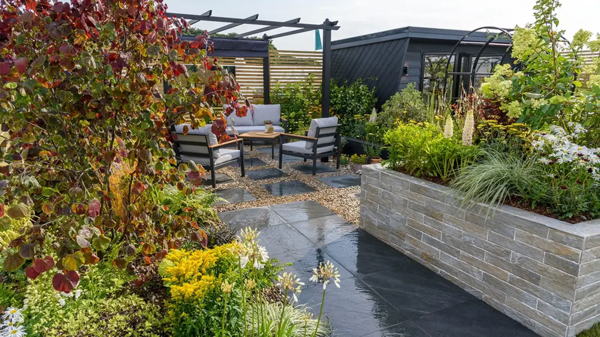 A modern garden has a black office cabin, a pergola over lounge seating, dark grey paving, and a raised stone planter filled with colourful plants.