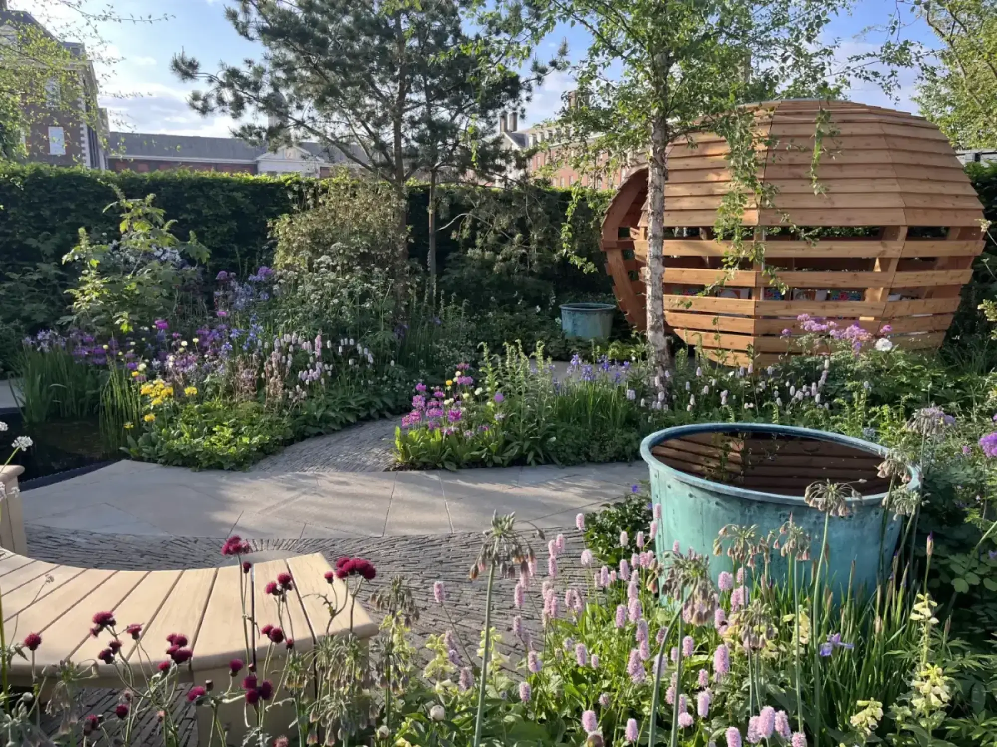 A small show garden with a large wooden spherical den, a curved timber bench, verdigris water tubs, and naturalistic planting.