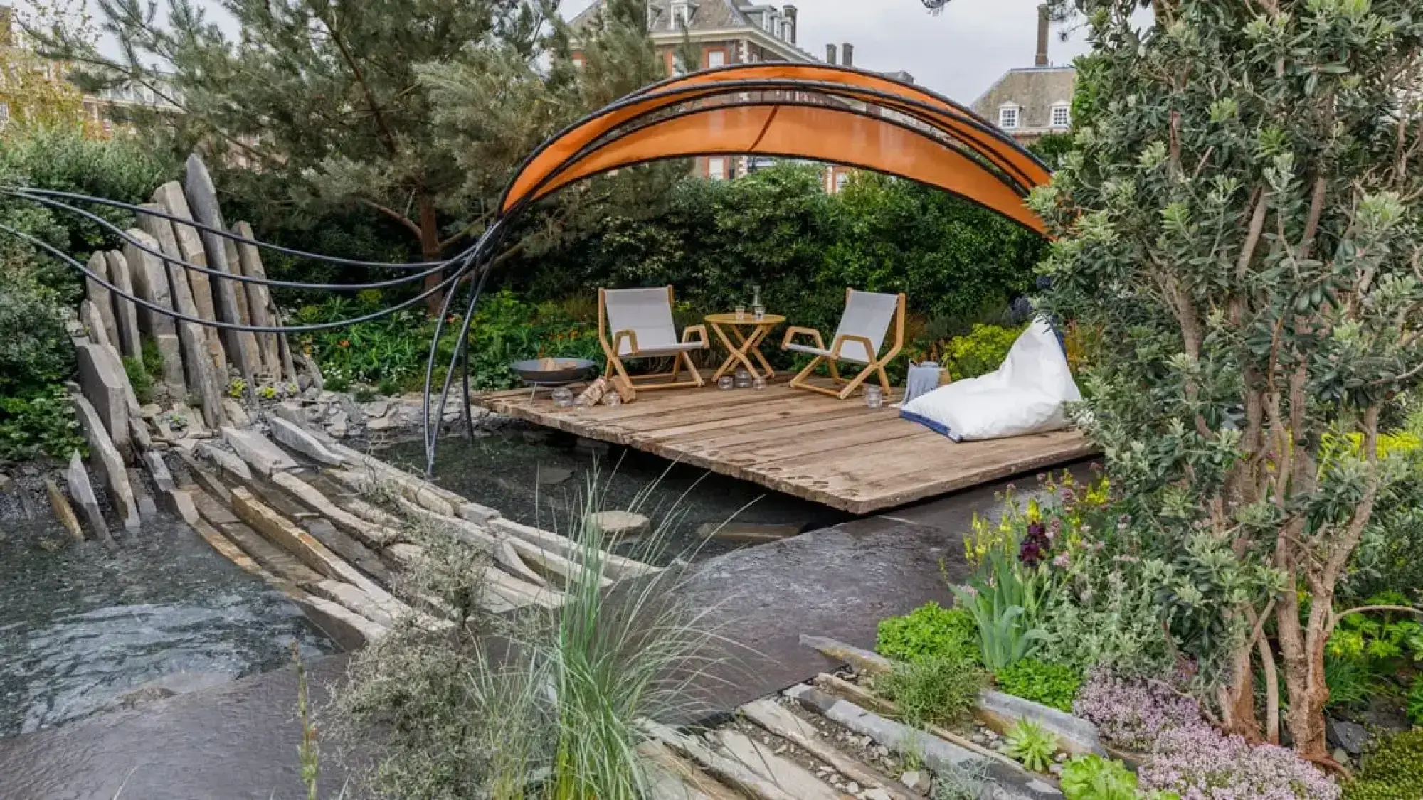 A contemporary garden featuring an orange fabric canopy over a timber deck with chairs and a fire pit, next to a slate waterfall into a pond.