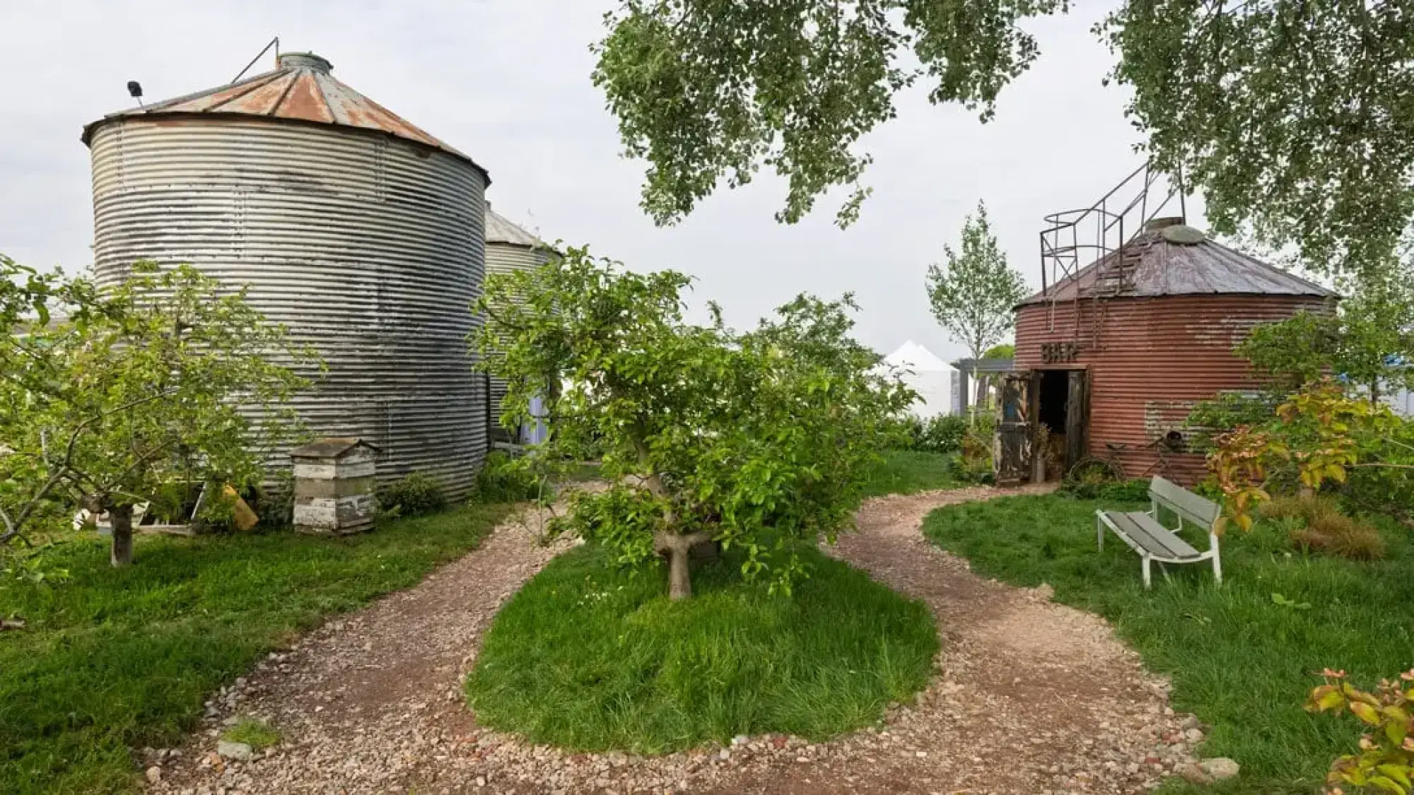 Two corrugated metal grain silos, one silver and one rusted red, stand in a garden with gravel paths, green grass, small fruit trees, a pale green bench, and a rustic beehive.