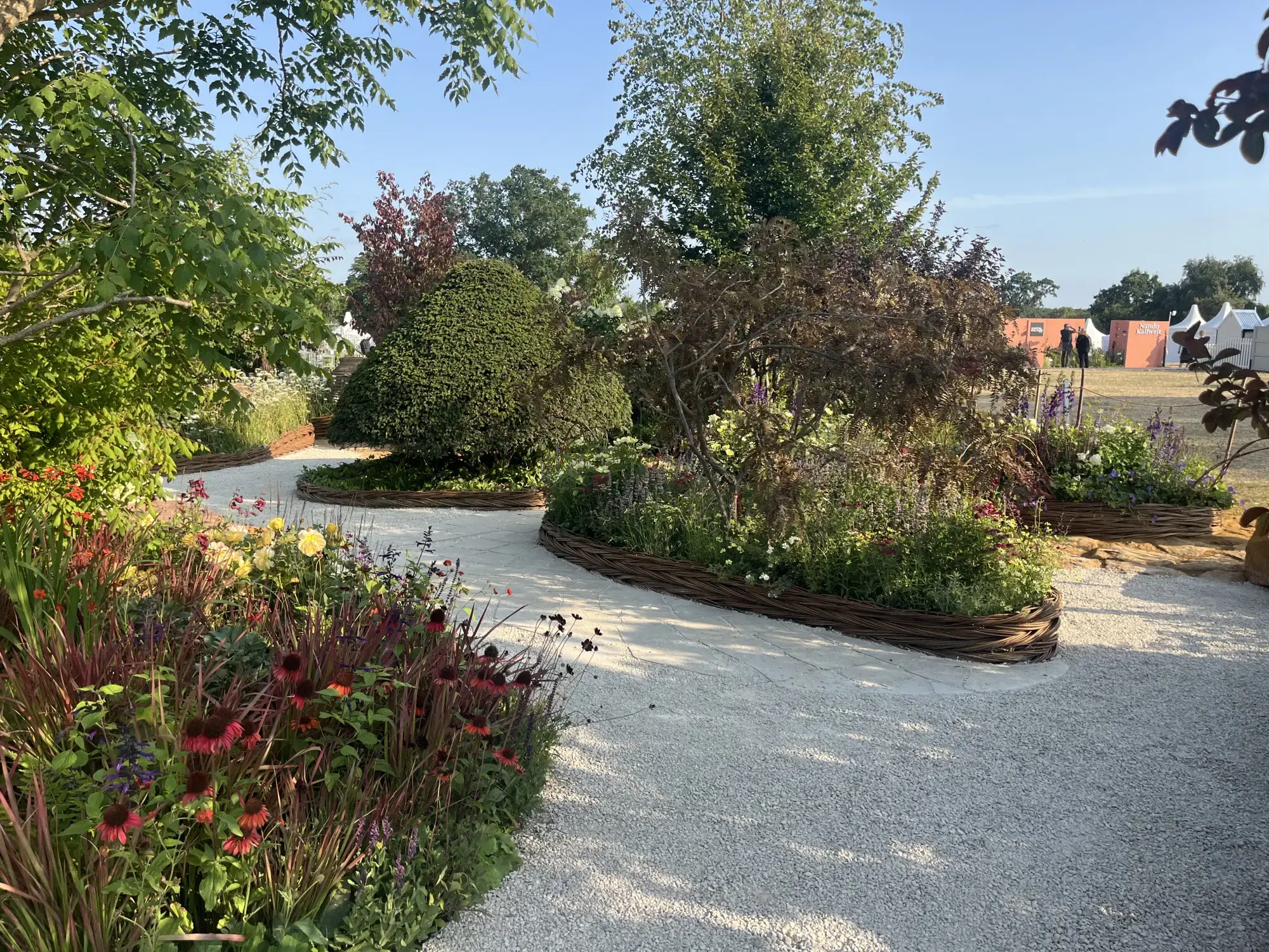 A meandering light grey gravel path winds through a sunny feature garden, bordered by woven willow edging and vibrant mixed herbaceous planting including red echinacea.