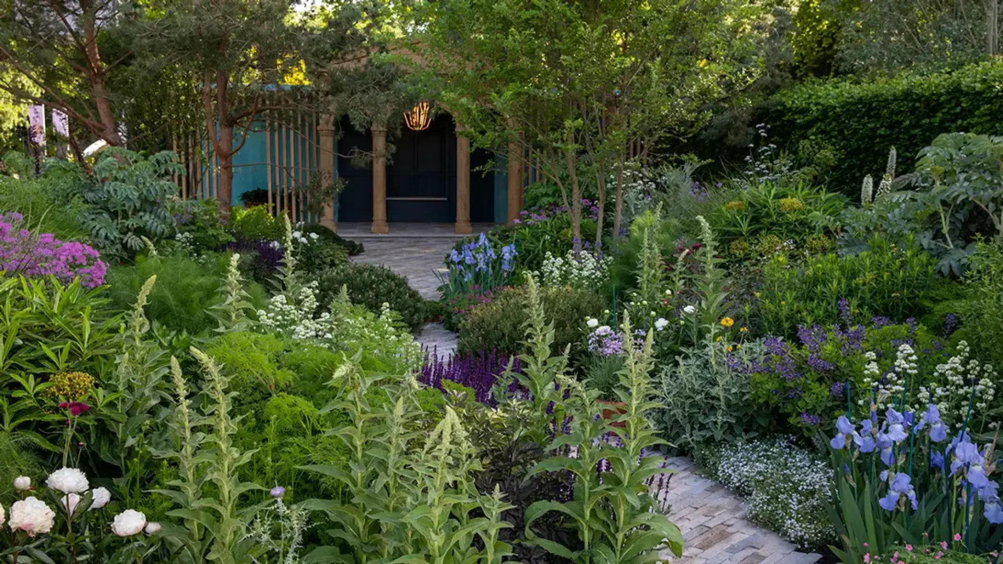 A naturalistic garden path winds past tall spires, blue irises, and purple flowers towards a dark blue boathouse entrance with timber columns.