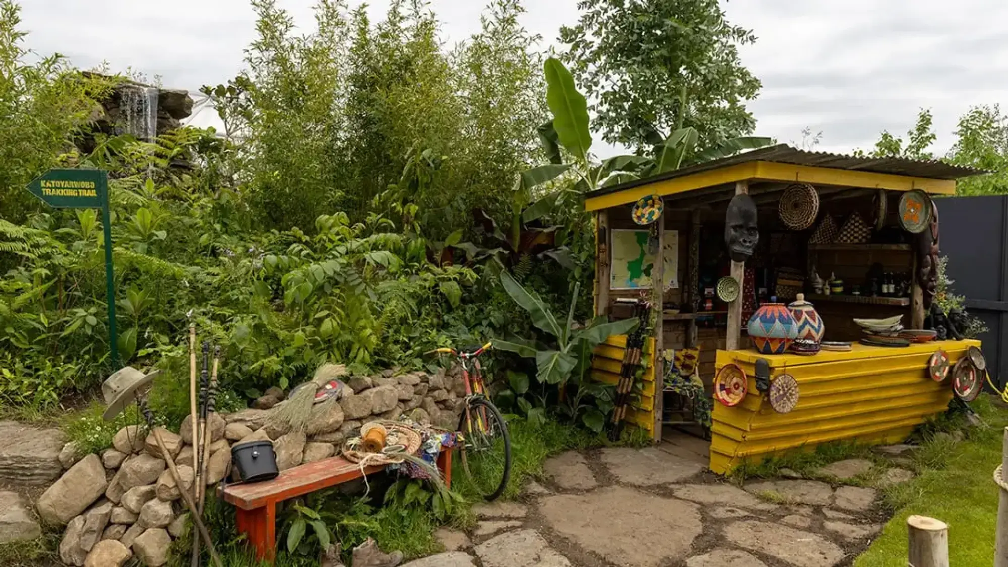 A vibrant, immersive show garden featuring a bright yellow corrugated metal shack, a rustic stone wall, a small waterfall, and lush tropical foliage with banana plants and ferns.