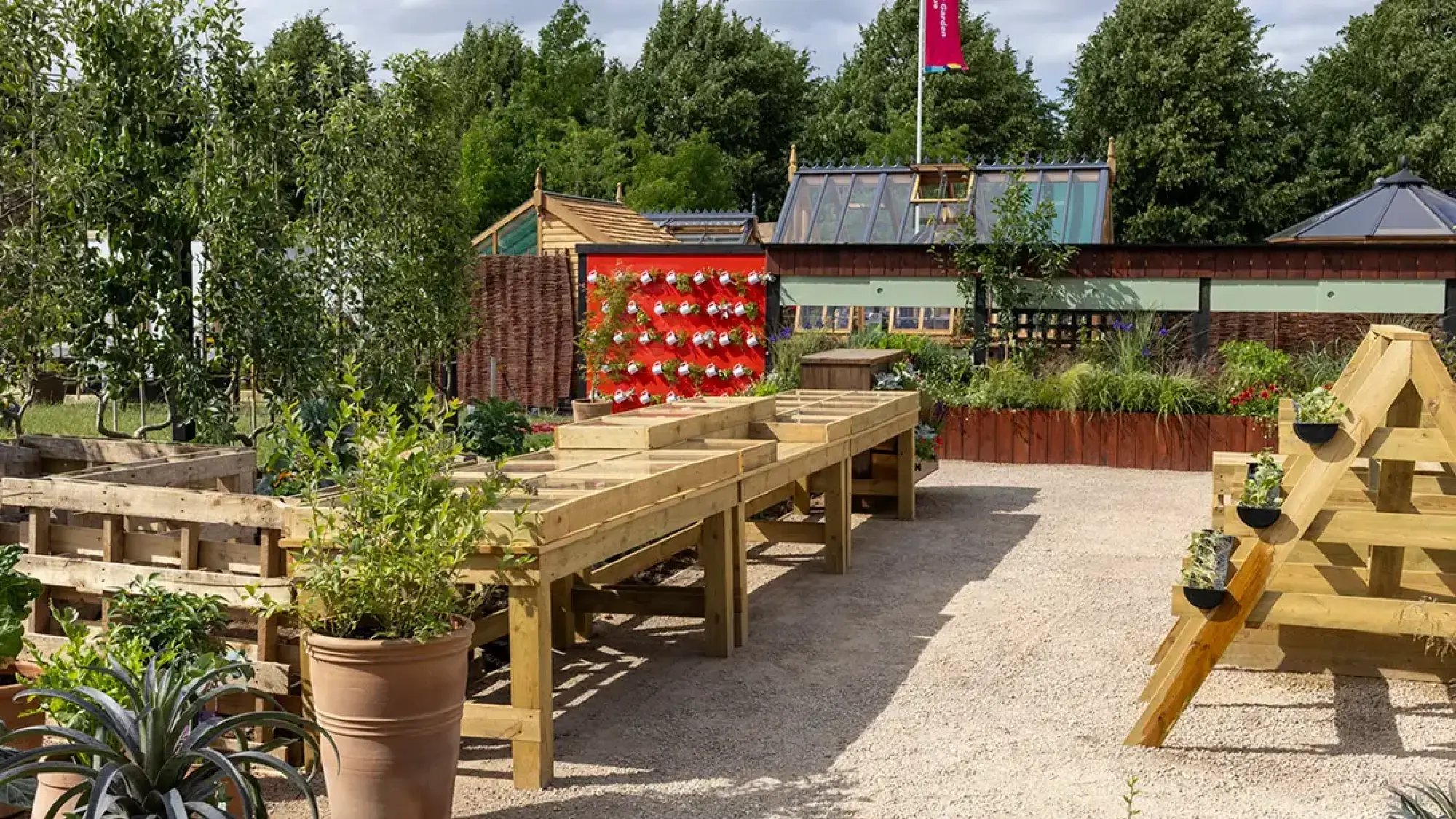A sunny, budget-friendly kitchen garden with a red vertical wall of watering can planters, wooden propagating tables, and pallet raised beds.