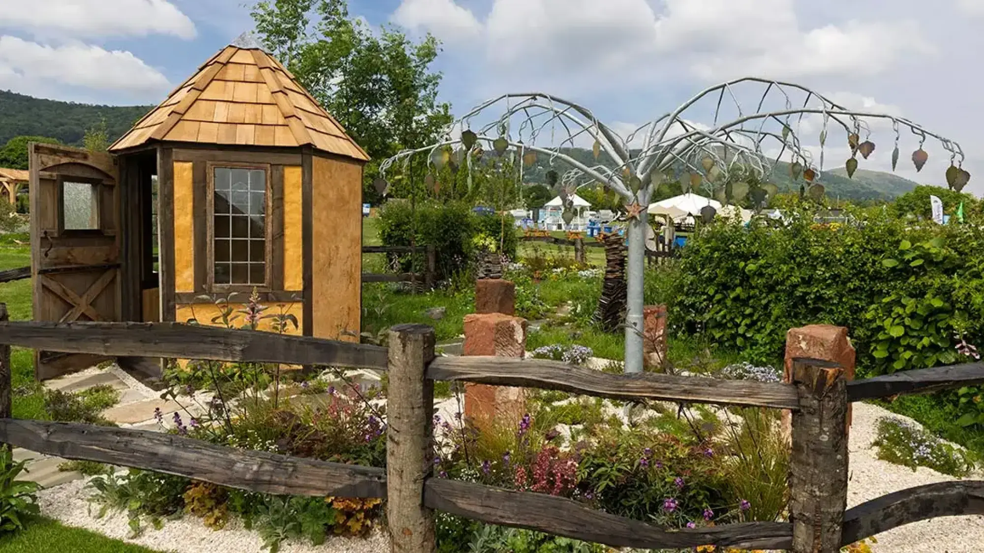 A rustic show garden features an octagonal timber hut, a metal tree sculpture, purple flowers, a gravel path, and a rough timber fence.