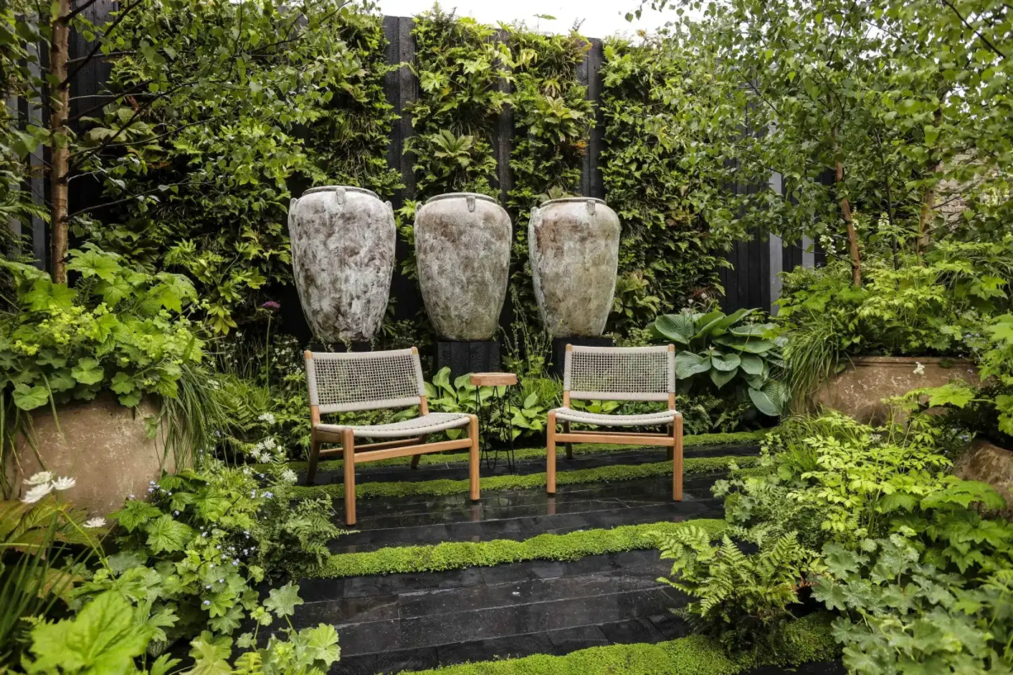 Three large aged urns against a dark timber living wall, surrounded by lush green foliage, black stepped paving, and two light wooden chairs.