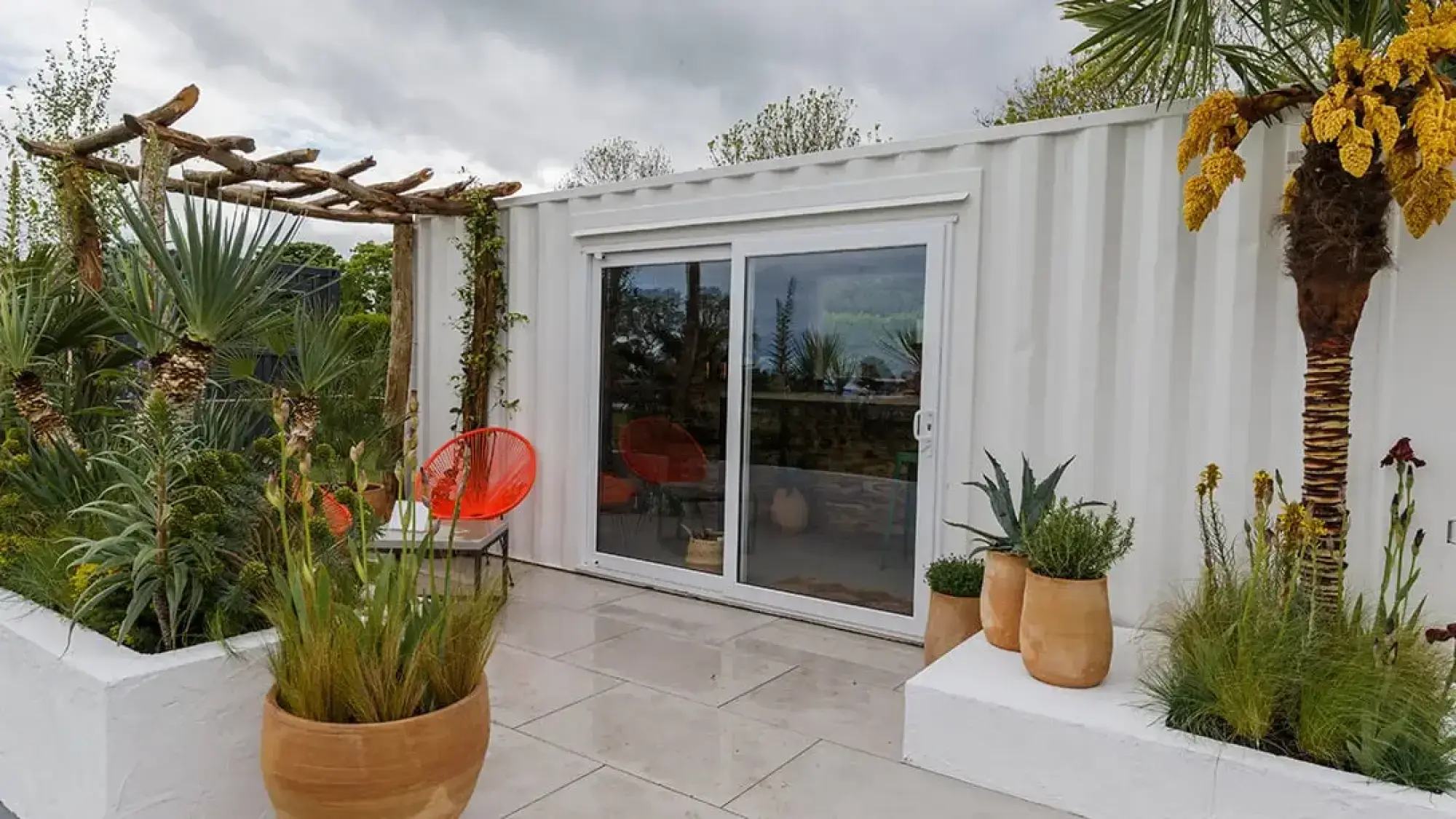 A Mediterranean terrace garden with a white container building, a rustic pergola, red chairs, and palms, agaves, and grasses in white raised beds and terracotta pots.