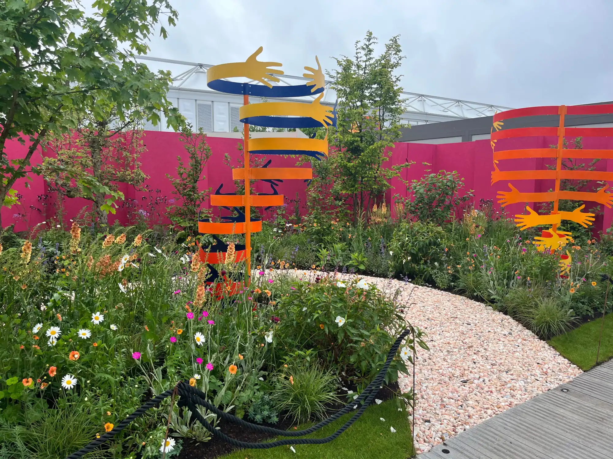 Vibrant garden with tall, colorful spiral hand sculptures against a bright pink wall, a pink gravel path, and mixed wildflowers.