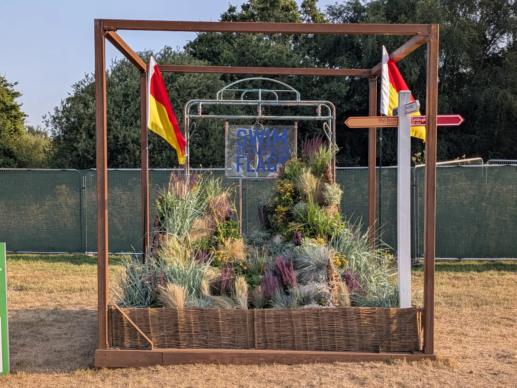 A beach-themed cube garden featuring a 'Swim Between the Flags' sign, lifeguard flags, grasses, and heather in a wicker planter.