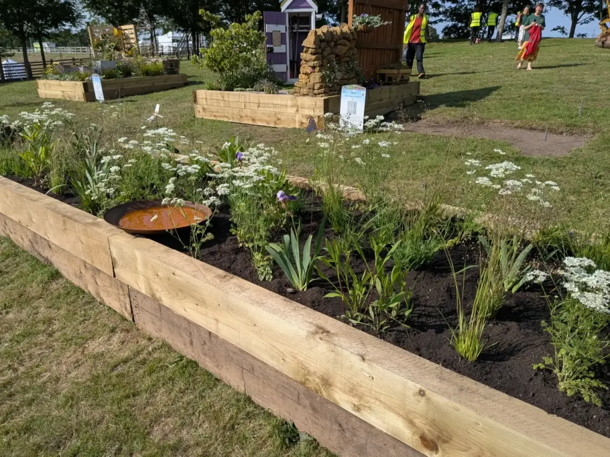 A raised wooden sleeper border garden features naturalistic planting with white and purple flowers, grasses, and a rust-coloured metal bird bath.