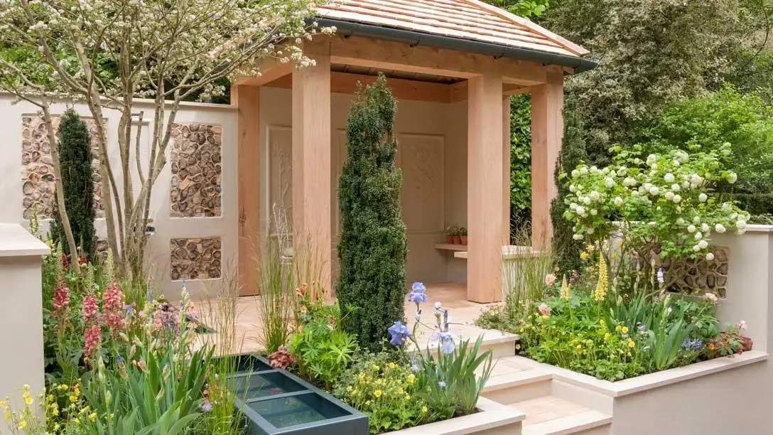 A tranquil Artisan Garden features a timber pavilion with a tiled roof, flint inlay walls, a reflective water feature, and steps leading through colorful mixed perennial plantings.