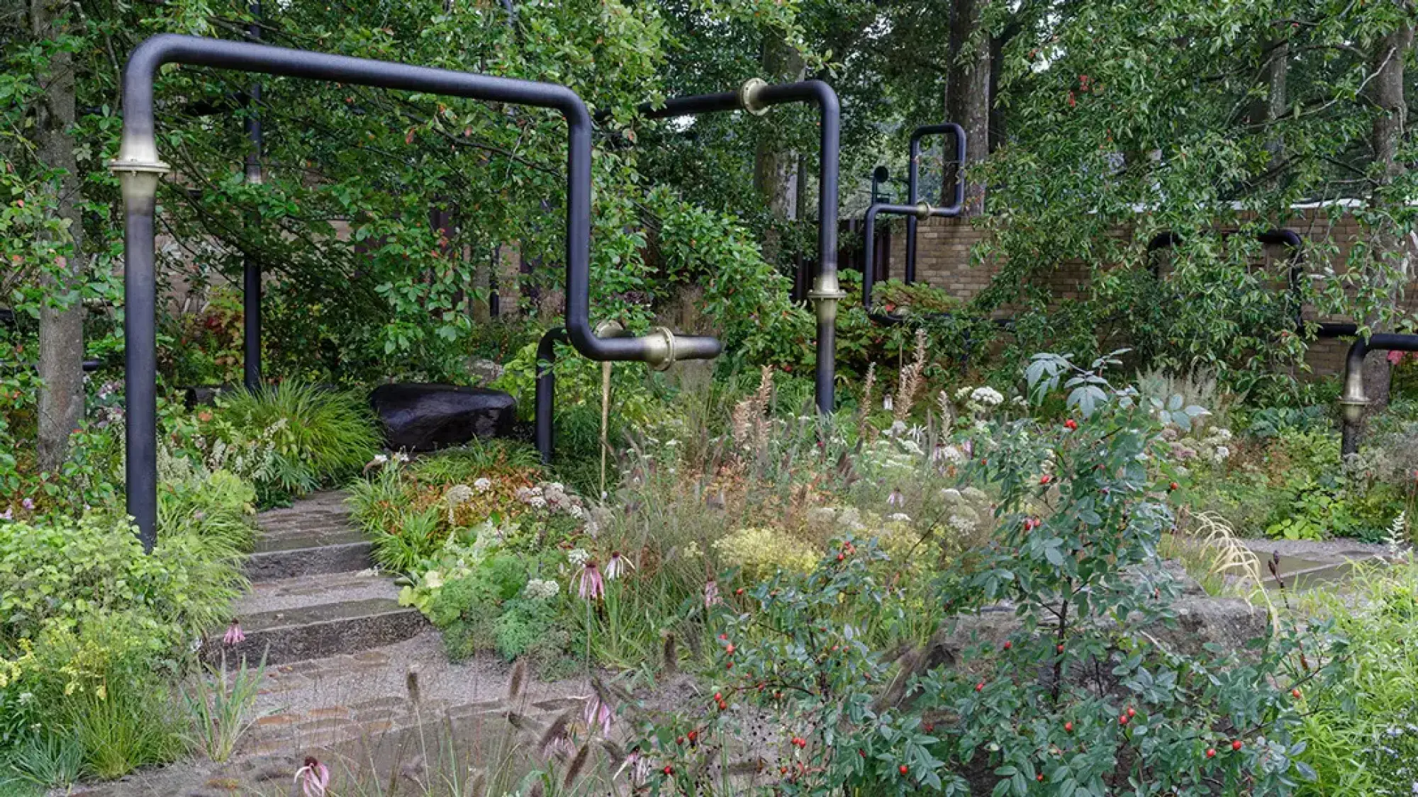 Black industrial pipes with brass fittings wind through a naturalistic garden with stone steps, a dark boulder, and mixed perennials.