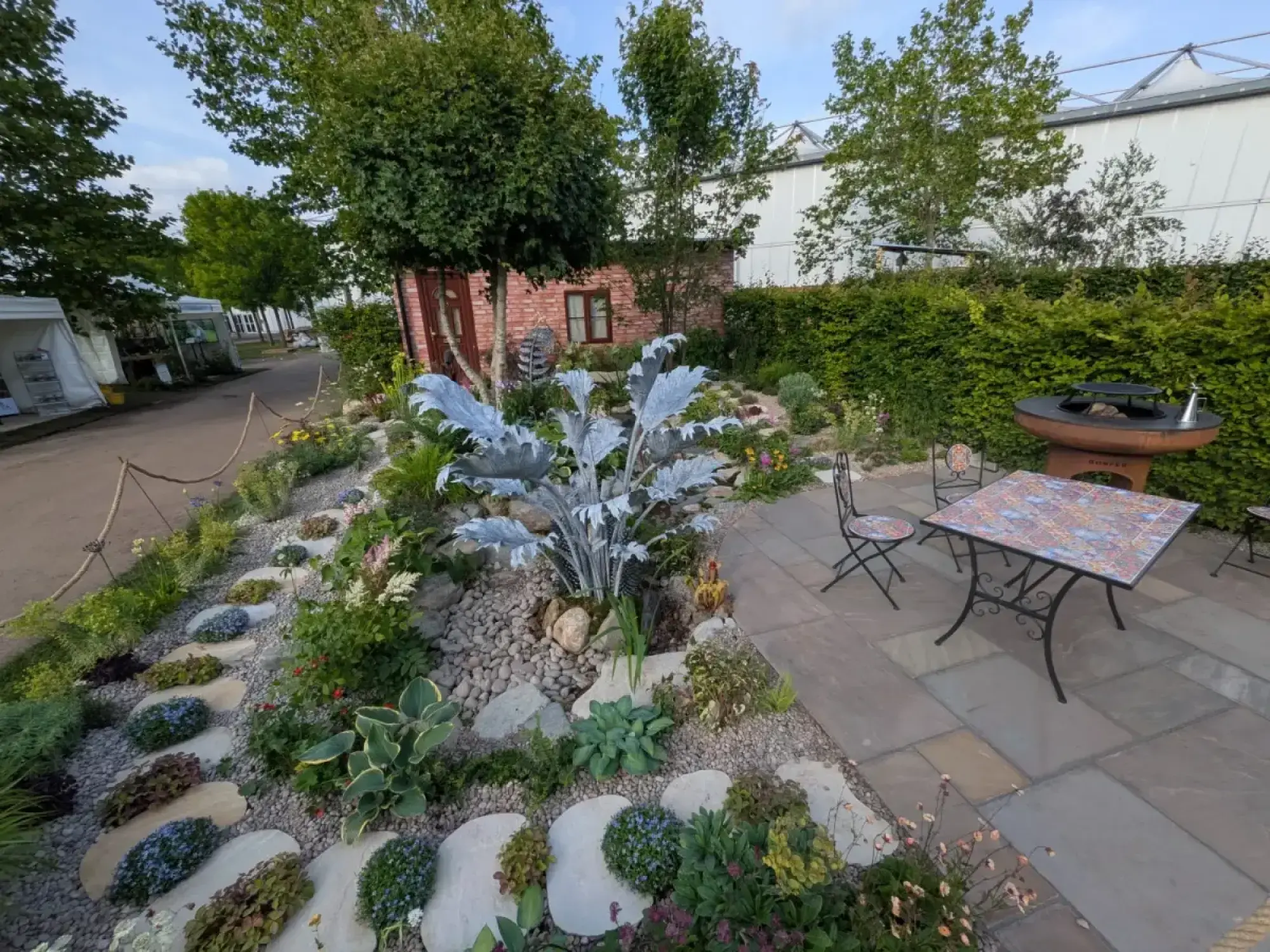 A contemporary garden features a large silver plant sculpture in a gravel bed with stepping stones, a paved patio with a mosaic table and fire pit, and a brick garden room at the back.