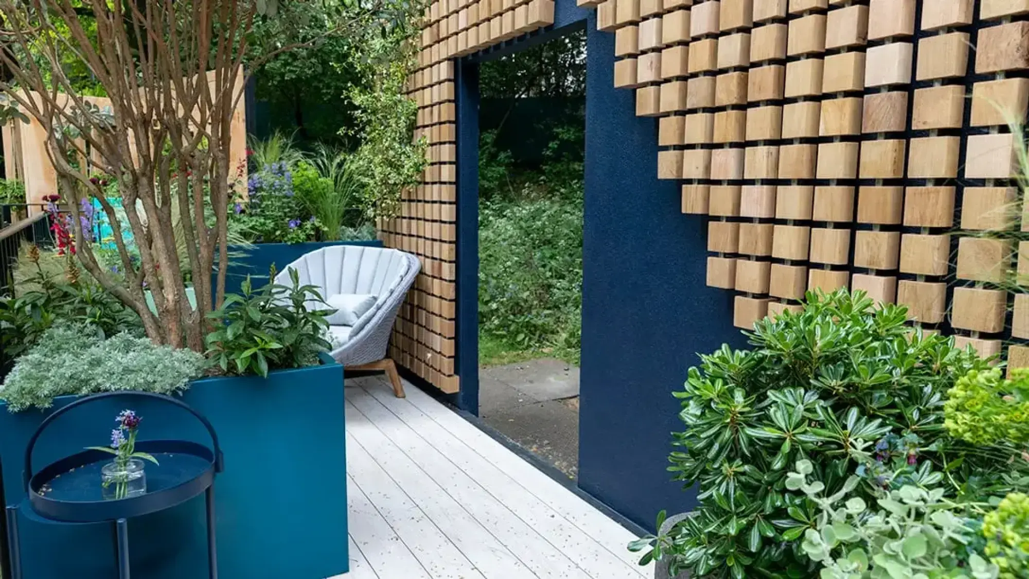 A contemporary balcony garden with a dark blue wall, wooden block mosaic, white decking, teal planters, a grey armchair, and diverse greenery.