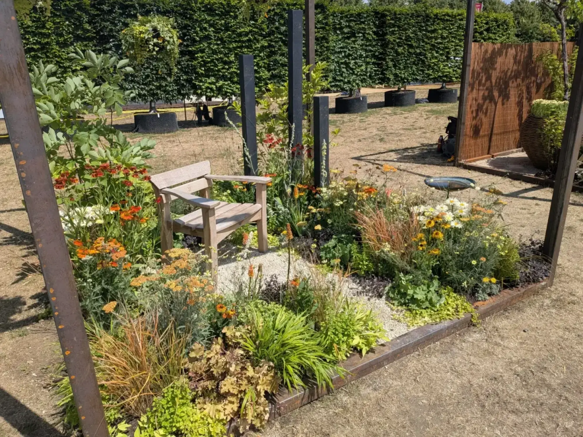 A cube garden features a corten steel frame, a wooden bench, and vibrant orange, yellow, and chartreuse flowers over light gravel.