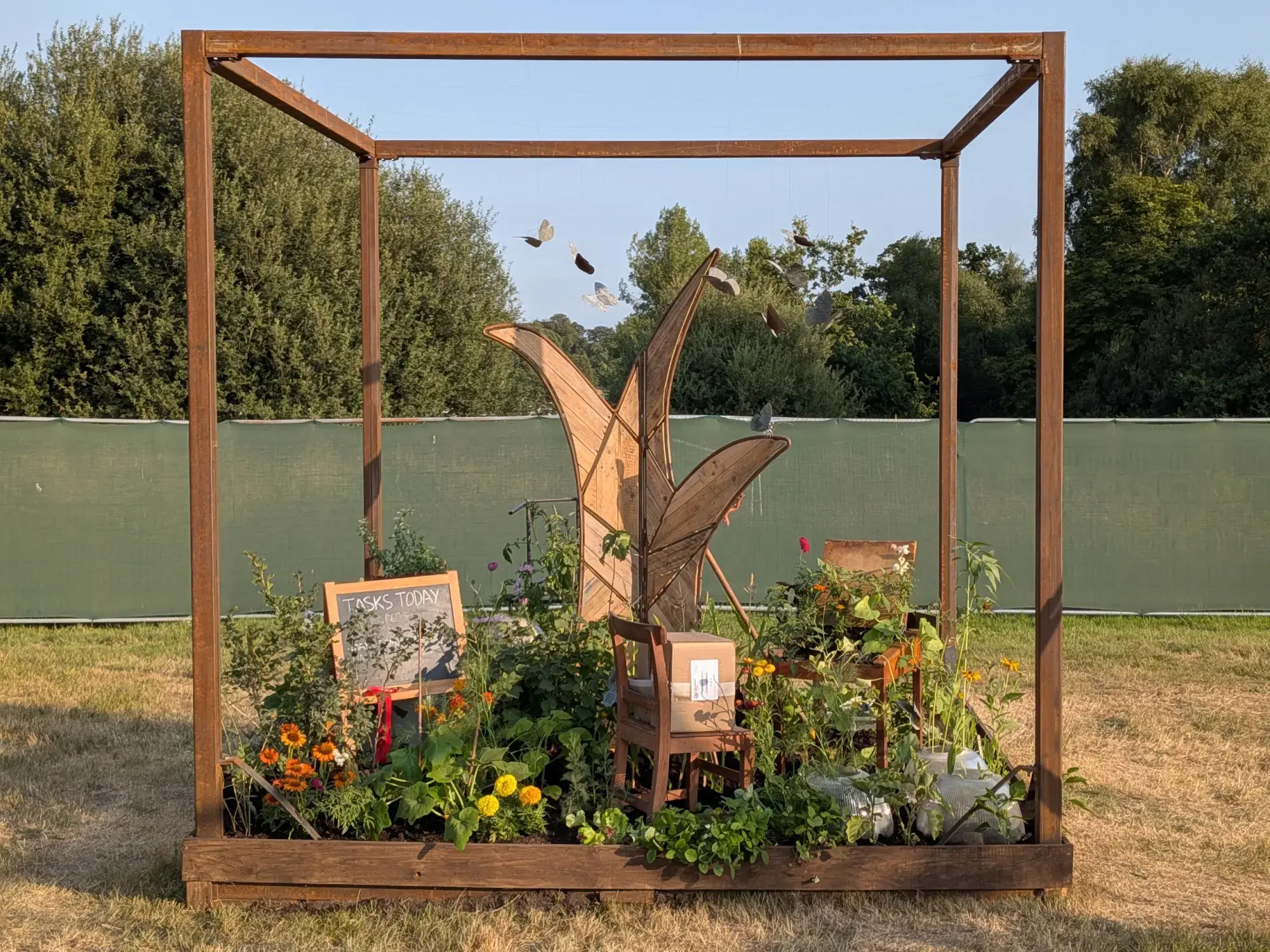 A rustic cube garden enclosed by a wooden frame, featuring an abstract wood sculpture, a butterfly mobile, chairs, and informal plantings of orange and yellow flowers.