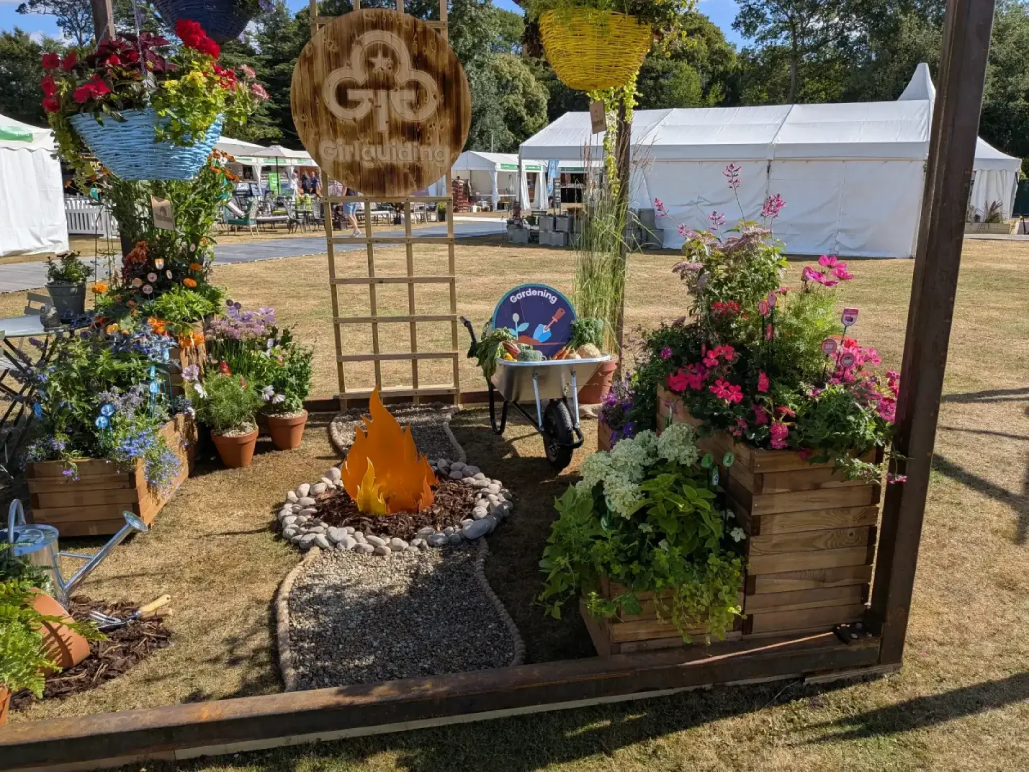 A cheerful cube garden features a faux campfire, vibrant flowers in wooden planters, a wheelbarrow with vegetables, and a Girlguiding sign.