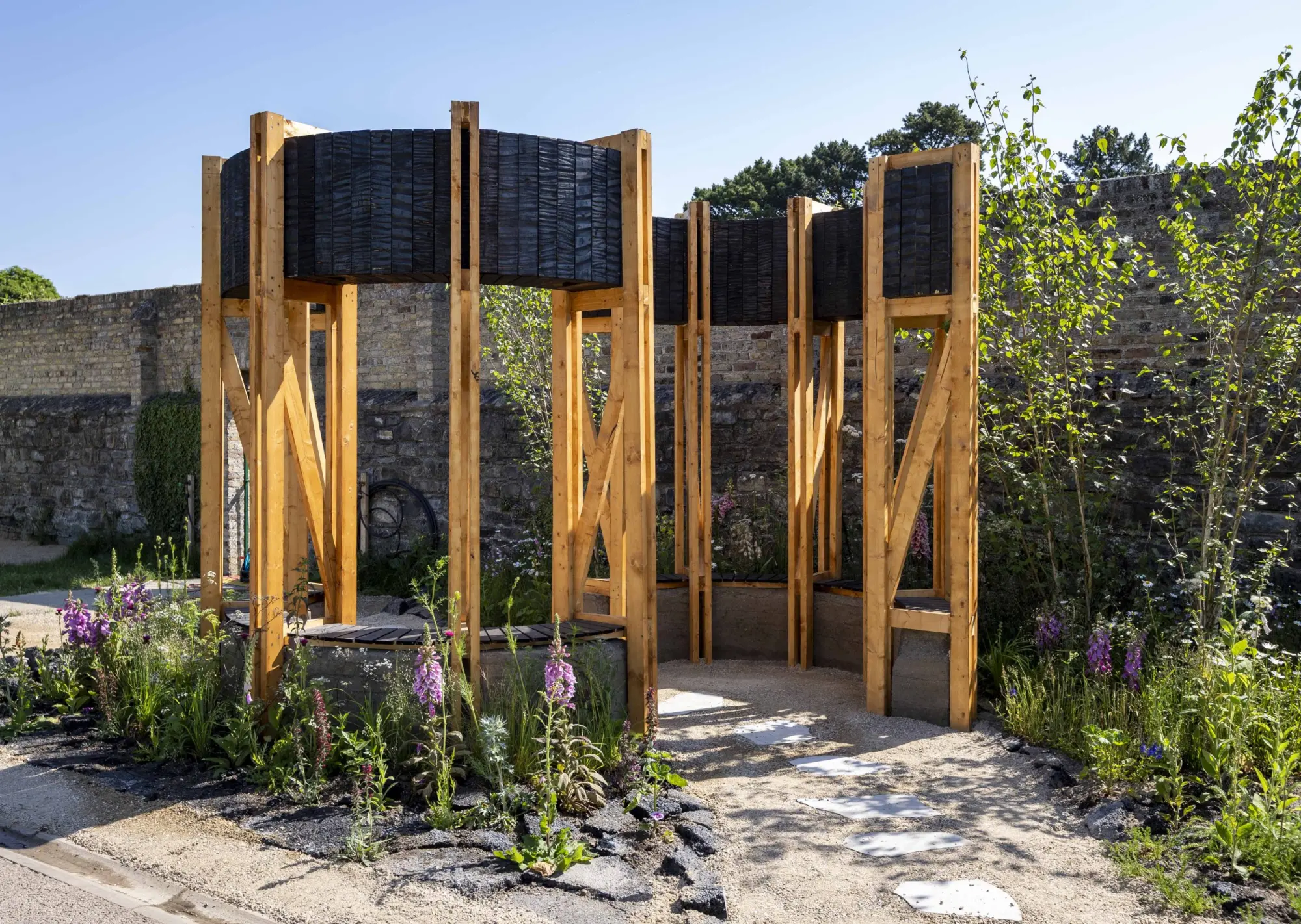 A modern concept garden features a circular light timber pavilion with dark charred wood cladding, an integrated bench, gravel path, and purple foxglove planting.