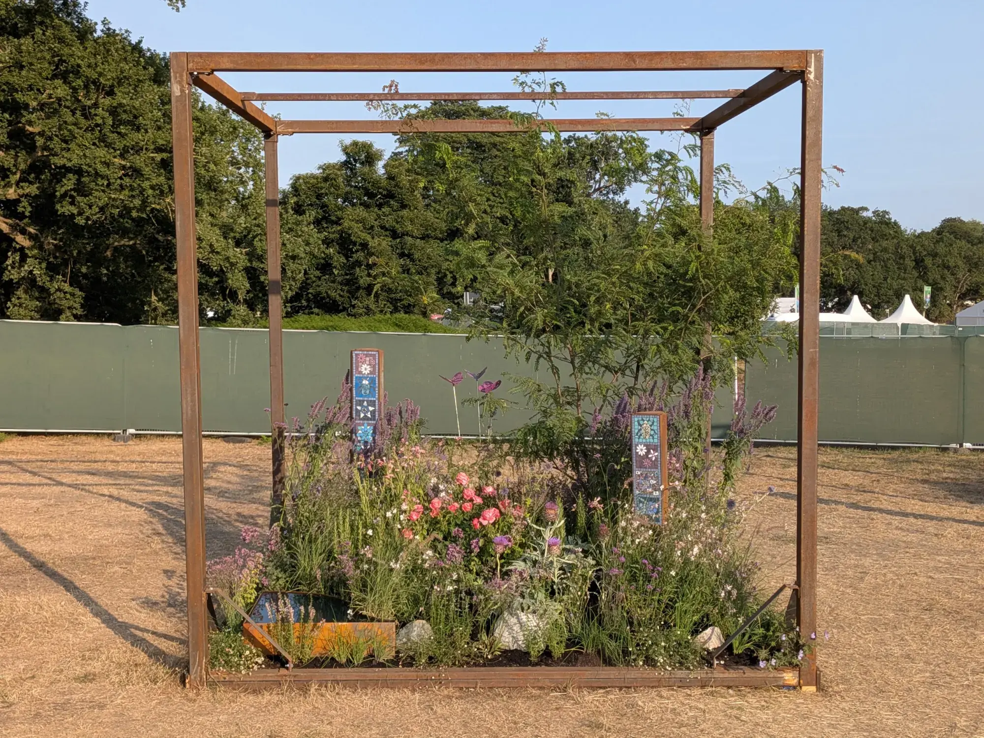 A rust metal cube frames a naturalistic garden with pink roses, purple flowers, grasses, a small tree, mosaic panels, and a reflective water element.