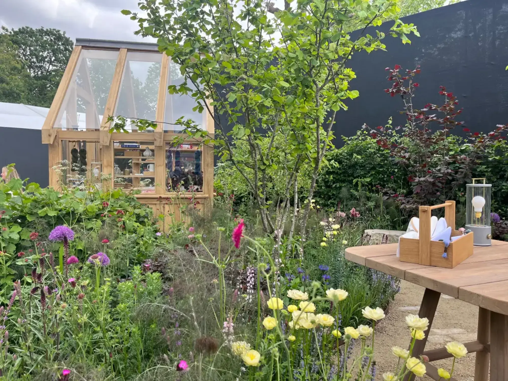 A vibrant garden showcases a wooden greenhouse, a timber picnic table, and flowering perennials in purple, yellow, and pink.