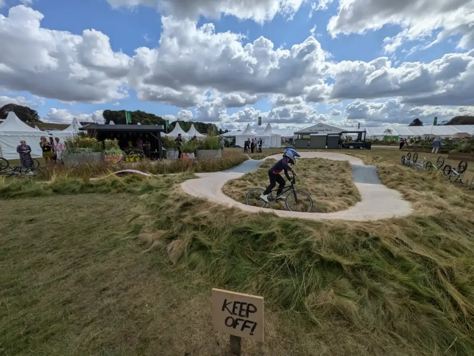 A concrete BMX pump track, surrounded by shaggy mounds of wild grass, is ridden by a person on a bicycle in a sunny feature garden.