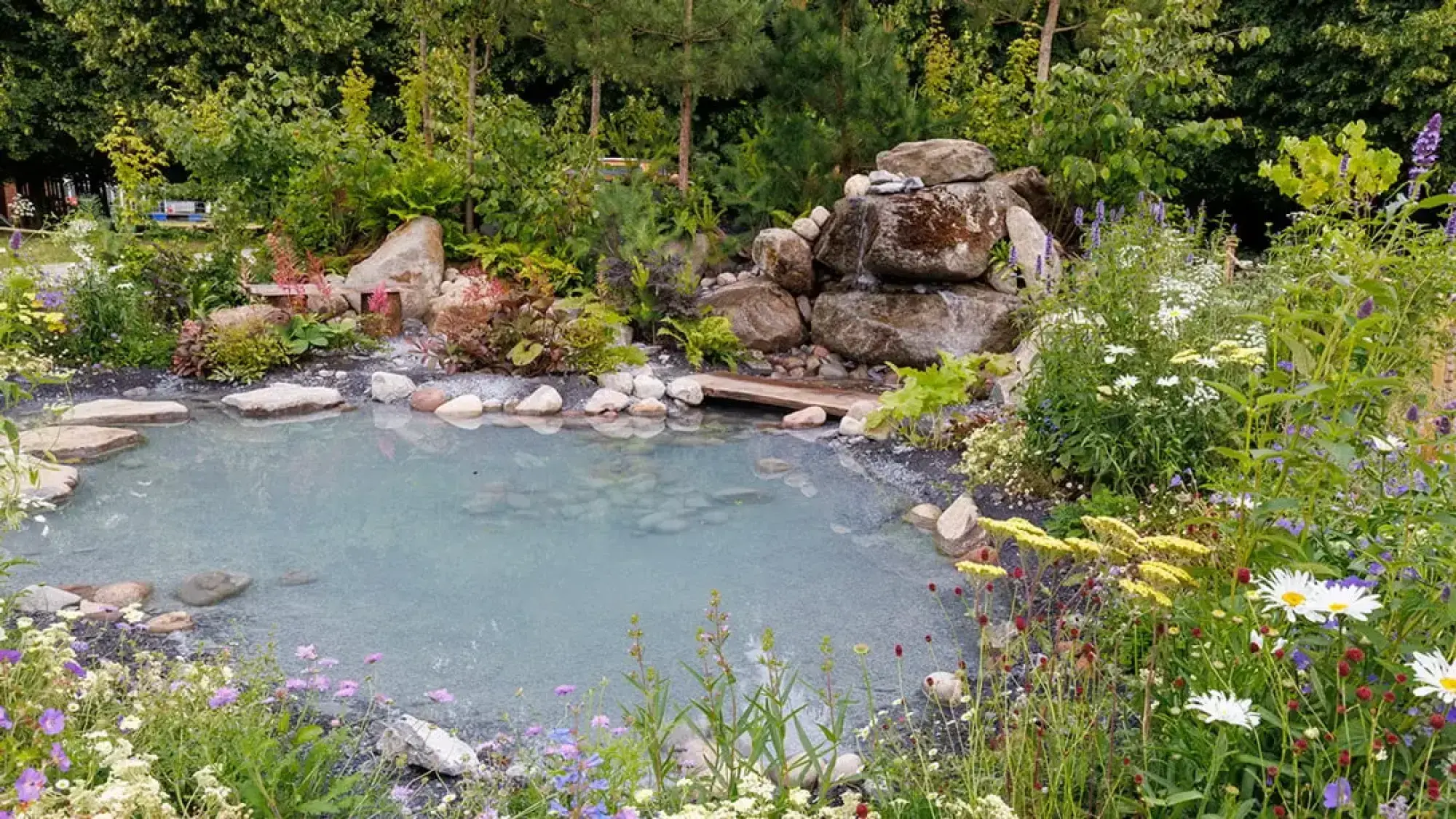 The Oregon Garden showcases a pale blue pond with stepping stones, a small wooden bridge, and a rock waterfall, set amongst lush, informal wildflowers.