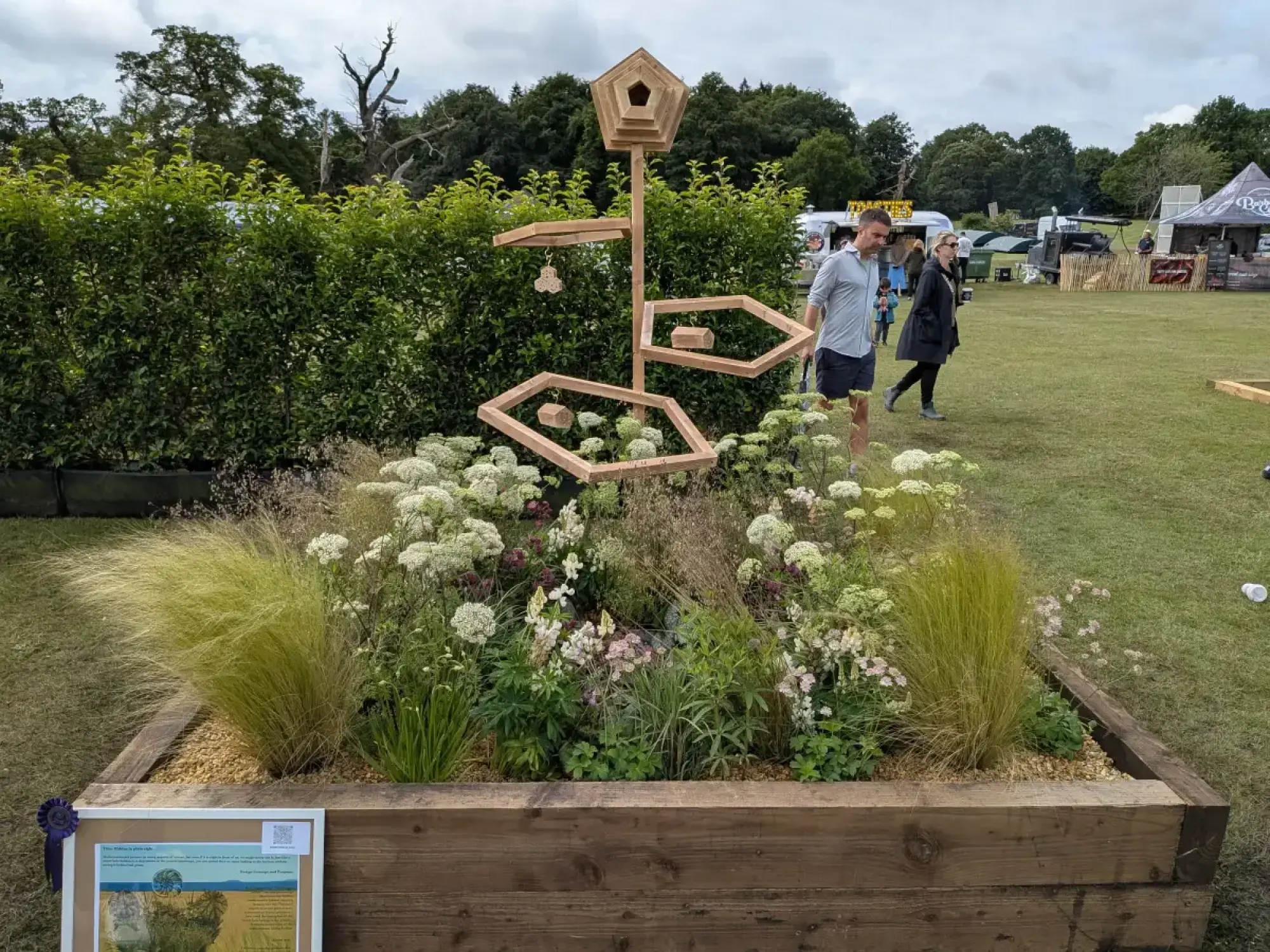 A raised timber bed holds naturalistic grasses and white flowers, with a tall wooden sculpture featuring a pentagonal birdhouse and hexagonal frames.