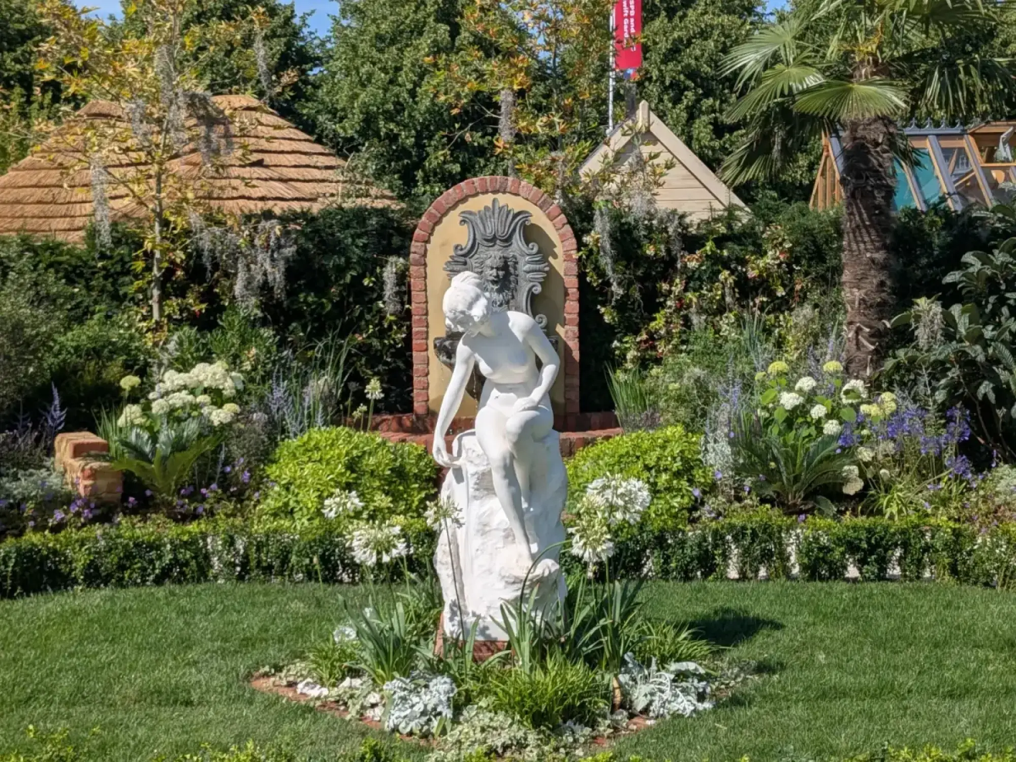 A show garden featuring a white classical statue in a central lawn, with lush mixed borders including a brick fountain, thatched roundhouse, timber shed, and a tall palm tree.