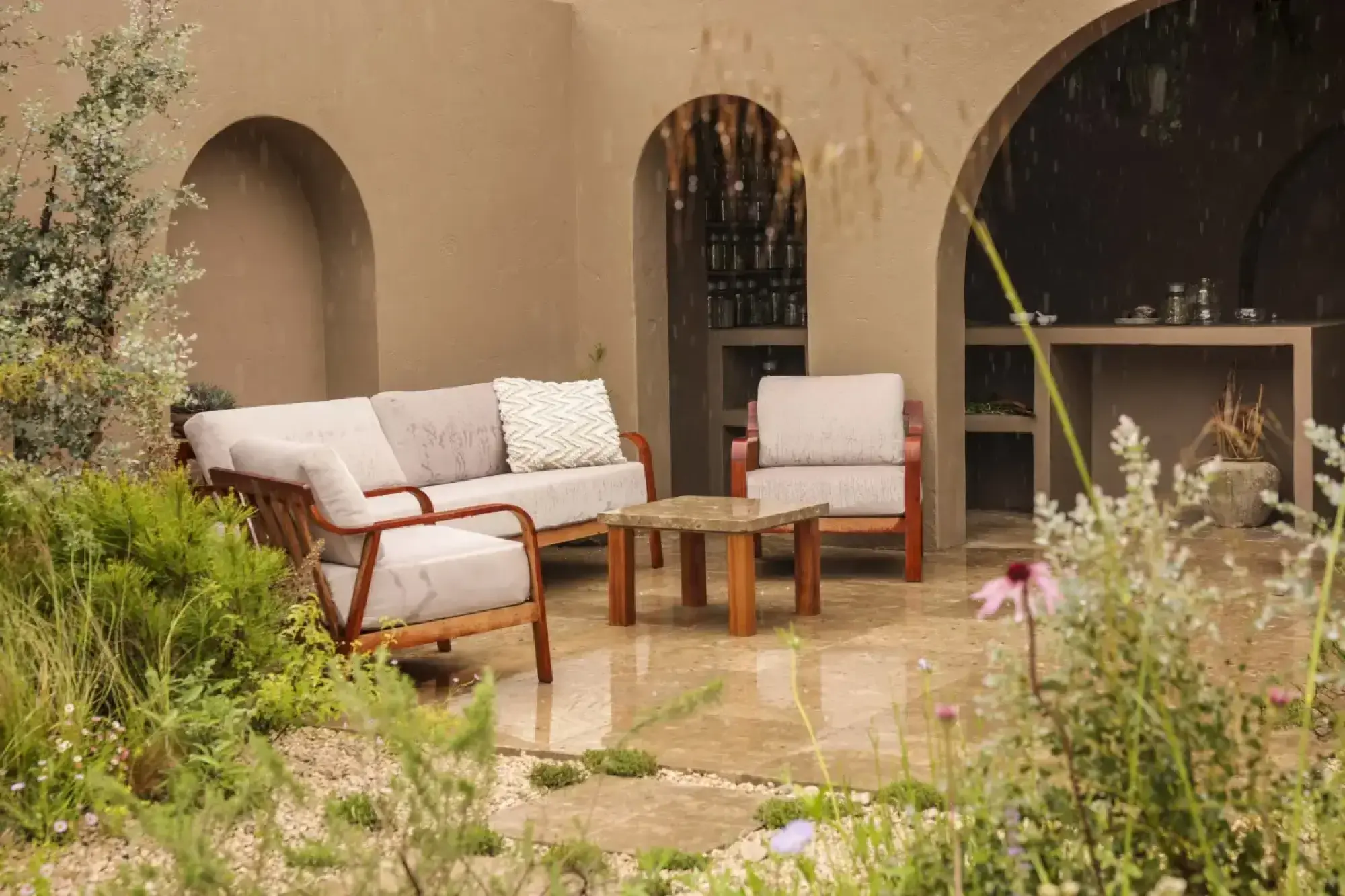 A tranquil modern rustic garden with a large flagstone patio, wooden lounge furniture, a rendered wall with arched alcoves, and naturalistic planting.