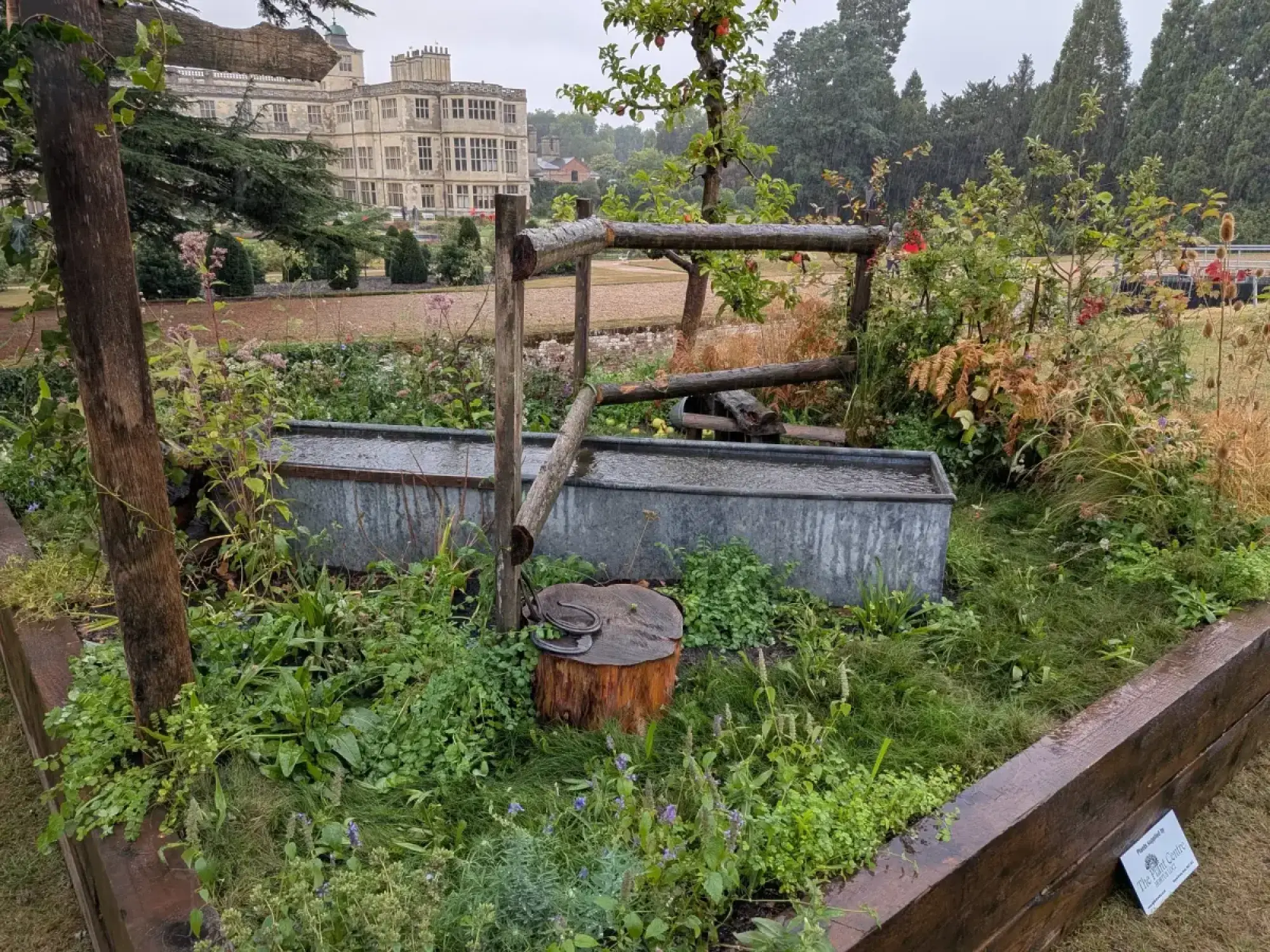 A naturalistic garden border featuring rustic wooden rails, two galvanised troughs, an apple tree, wild planting, and horseshoes on a tree stump.