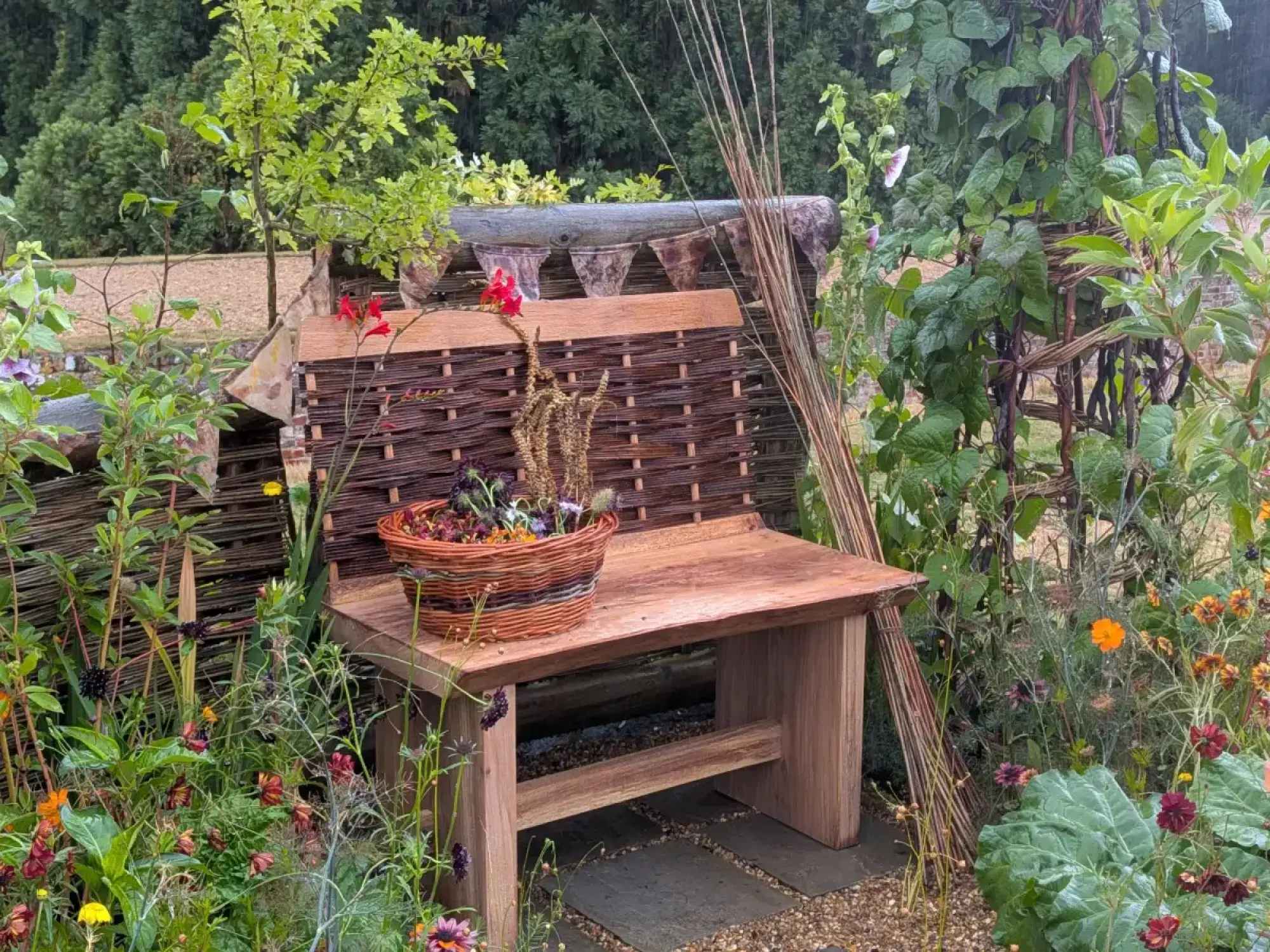 A rustic beautiful border featuring a woven willow bench, a woven fence, and a diverse mix of flowering plants in red, orange, purple, and green foliage.