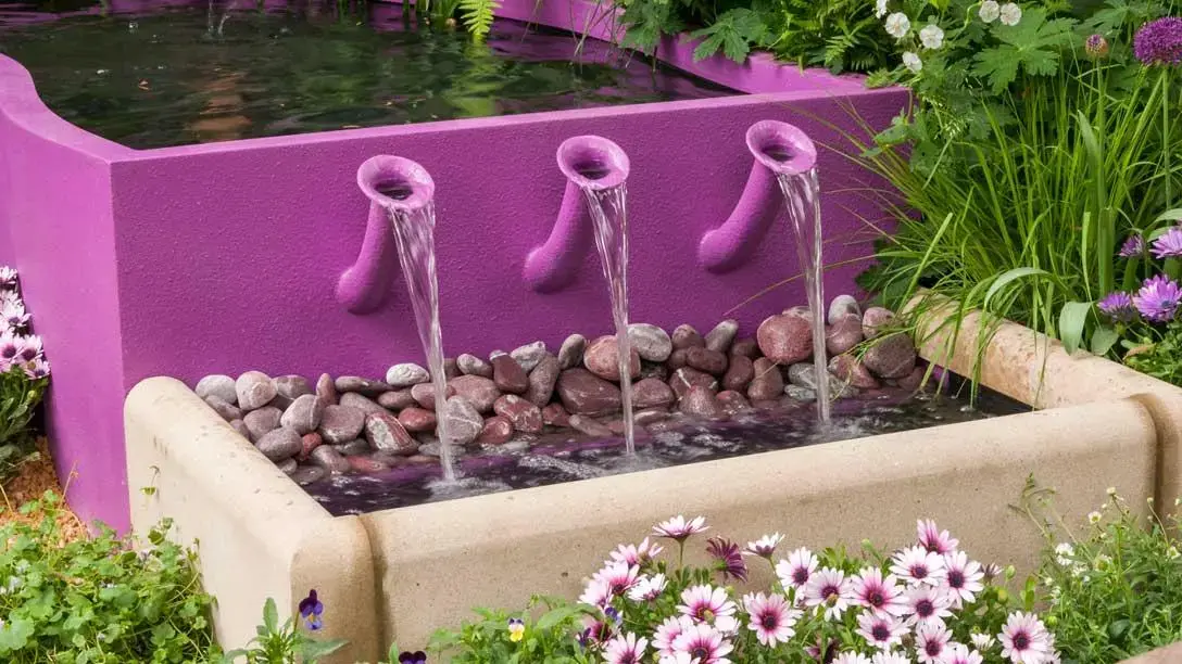 A bright magenta wall with three curved purple spouts cascades water into a beige stone trough filled with smooth river stones and surrounded by green plants and pink and purple flowers.