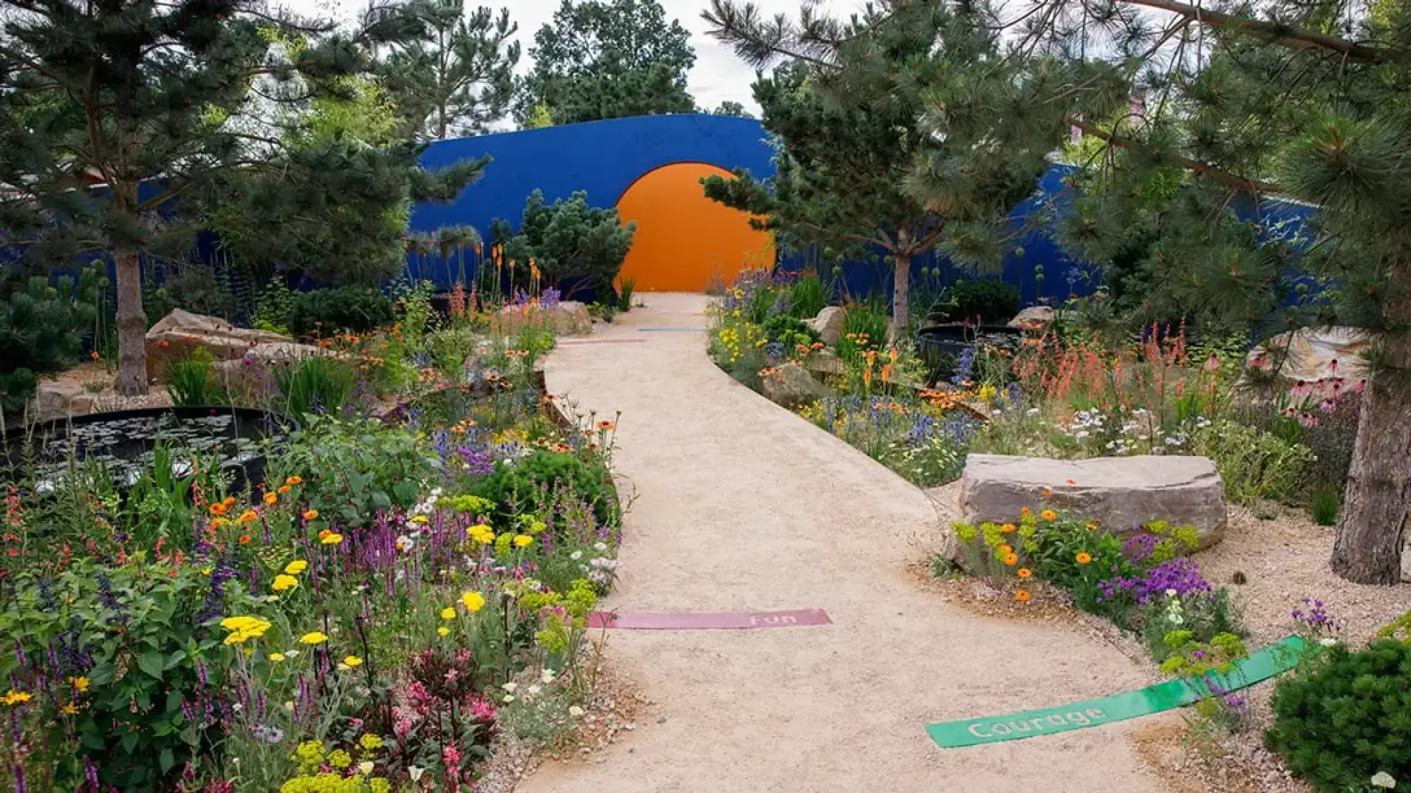 A winding gravel path leads through colourful herbaceous borders and pine trees towards a striking blue wall with a bright orange arched opening.