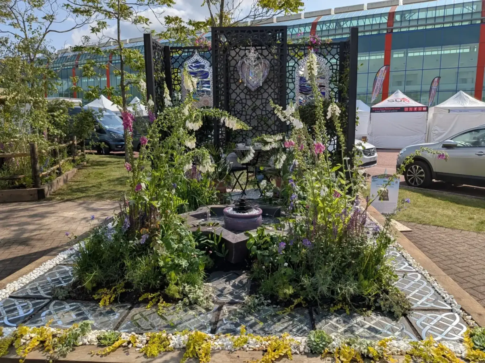 A sensory border garden with black screens, a heart mirror, central water feature, and white and pink foxgloves on Celtic knot paving.