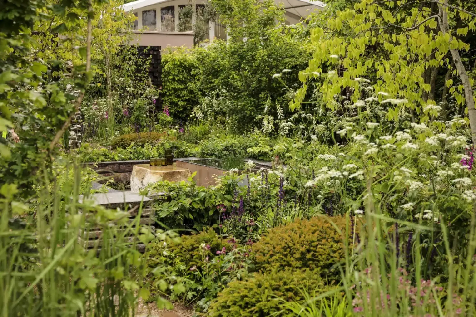 A naturalistic show garden features a stone water basin, a rectangular pool, a tall recycled bottle sculpture, and green and white plants.