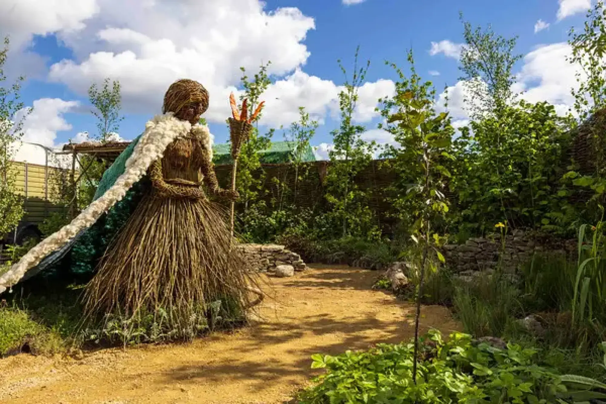 Woven Brigid sculpture in a green and white cloak stands on a gravel path, surrounded by young trees, green plants, and dry stone walls.
