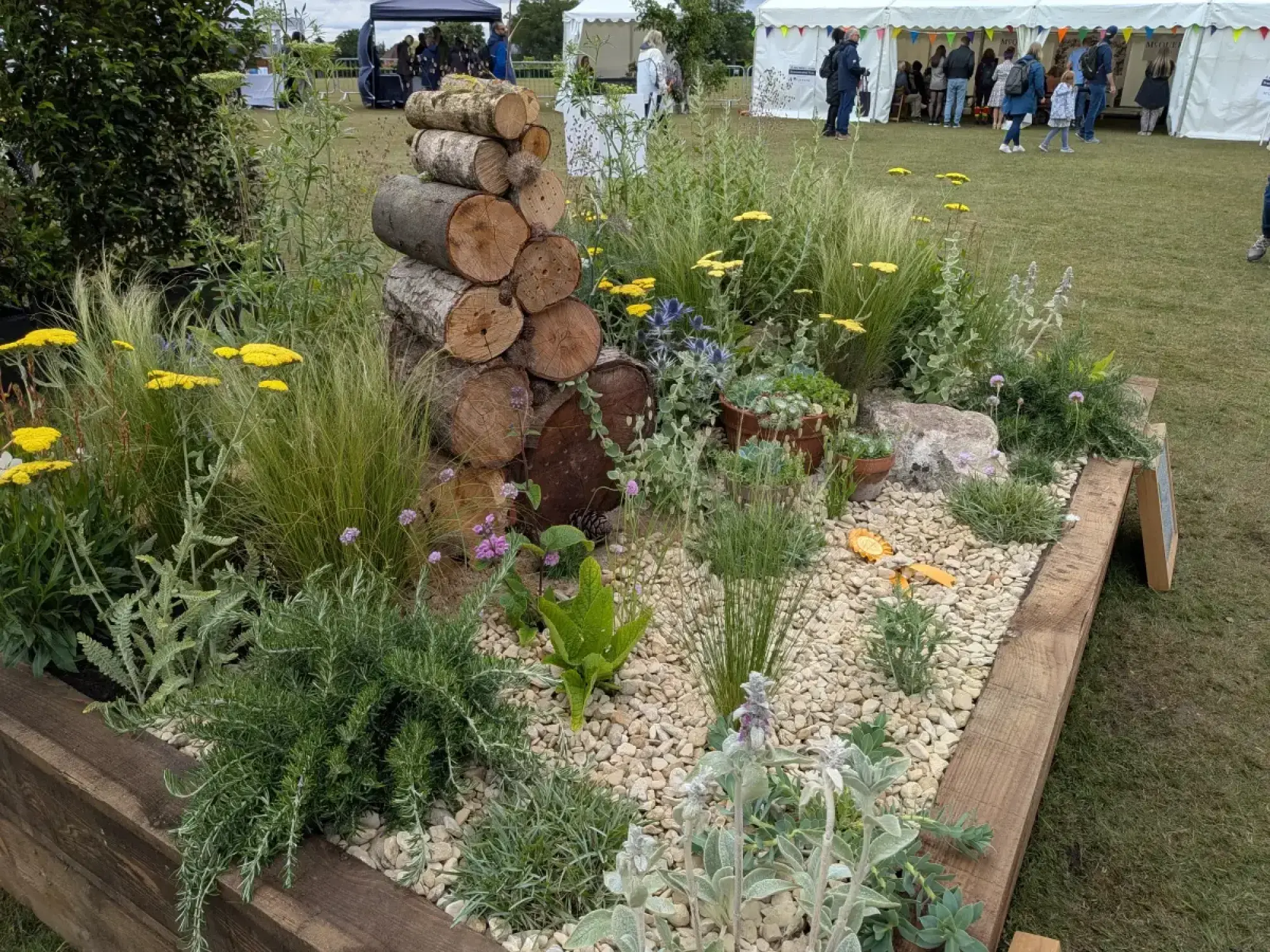 A raised garden bed features a stacked log pile, surrounded by ornamental grasses, yellow yarrow, purple flowers, and silver-leafed plants on a gravel surface.
