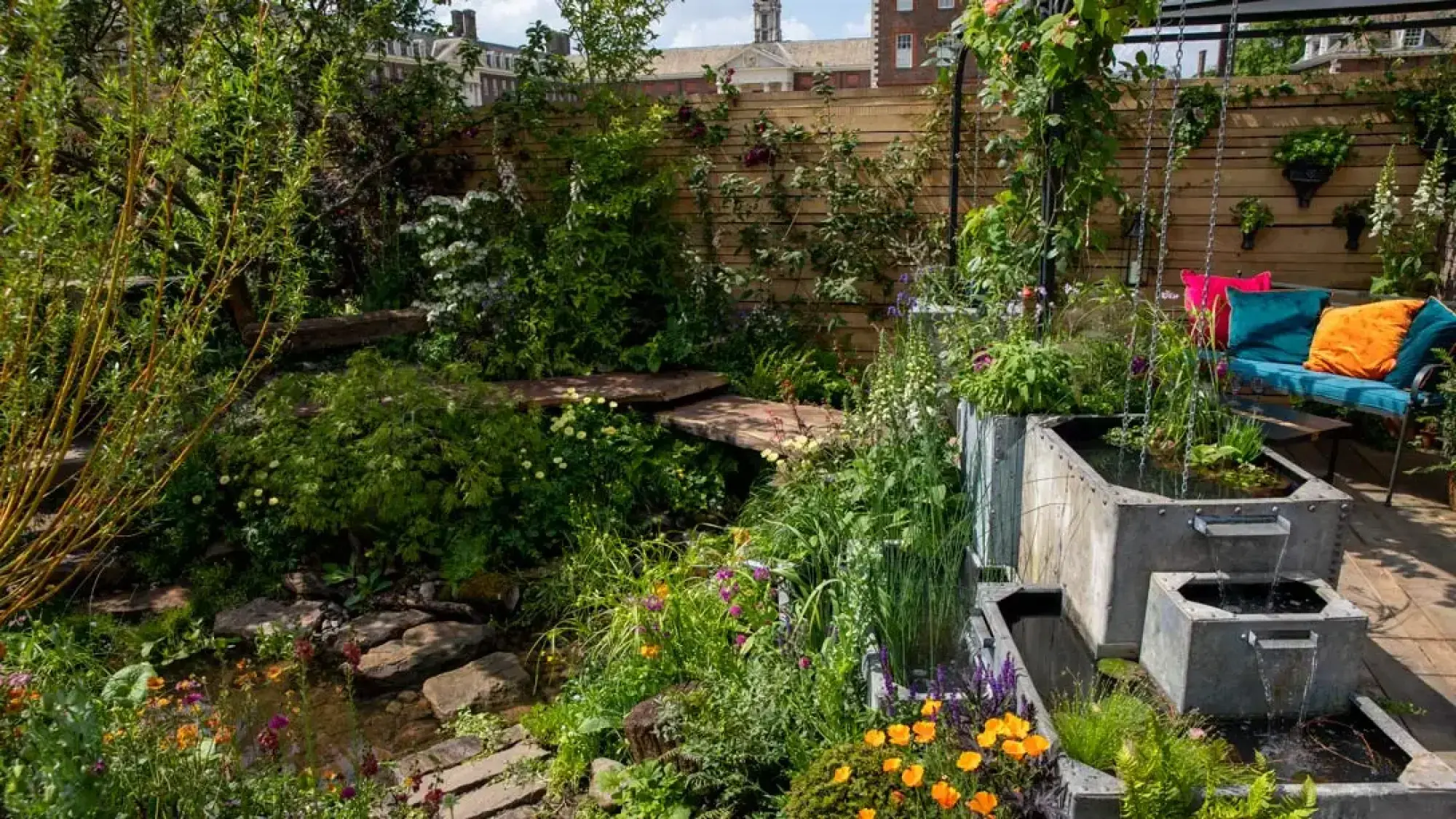 A tranquil, flood-resilient sanctuary garden with a tiered steel water feature, a stream, floating timber steps, and a blue sofa with cushions.
