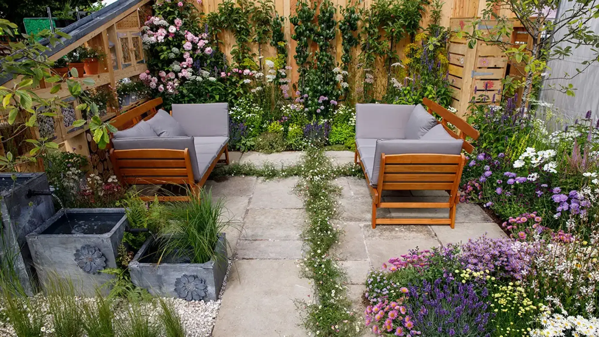 A vibrant small garden features a paved patio with a central daisy path, wooden sofas, a tiered water feature, and abundant wildflowers.