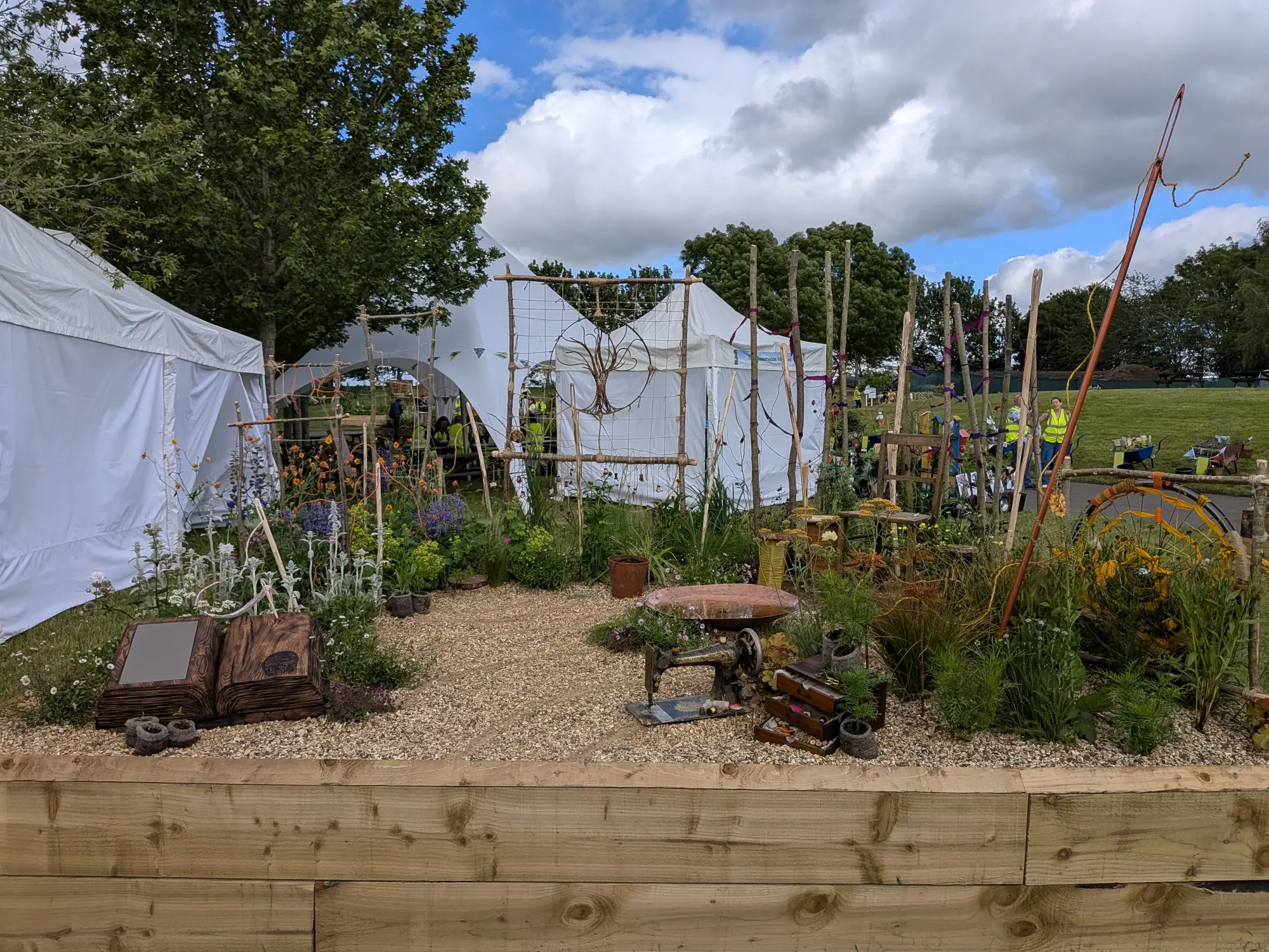 A 'Threads of Life' border features a tall sewing needle, a wire tree, an antique sewing machine, and an open wooden book among mixed planting and a gravel path.