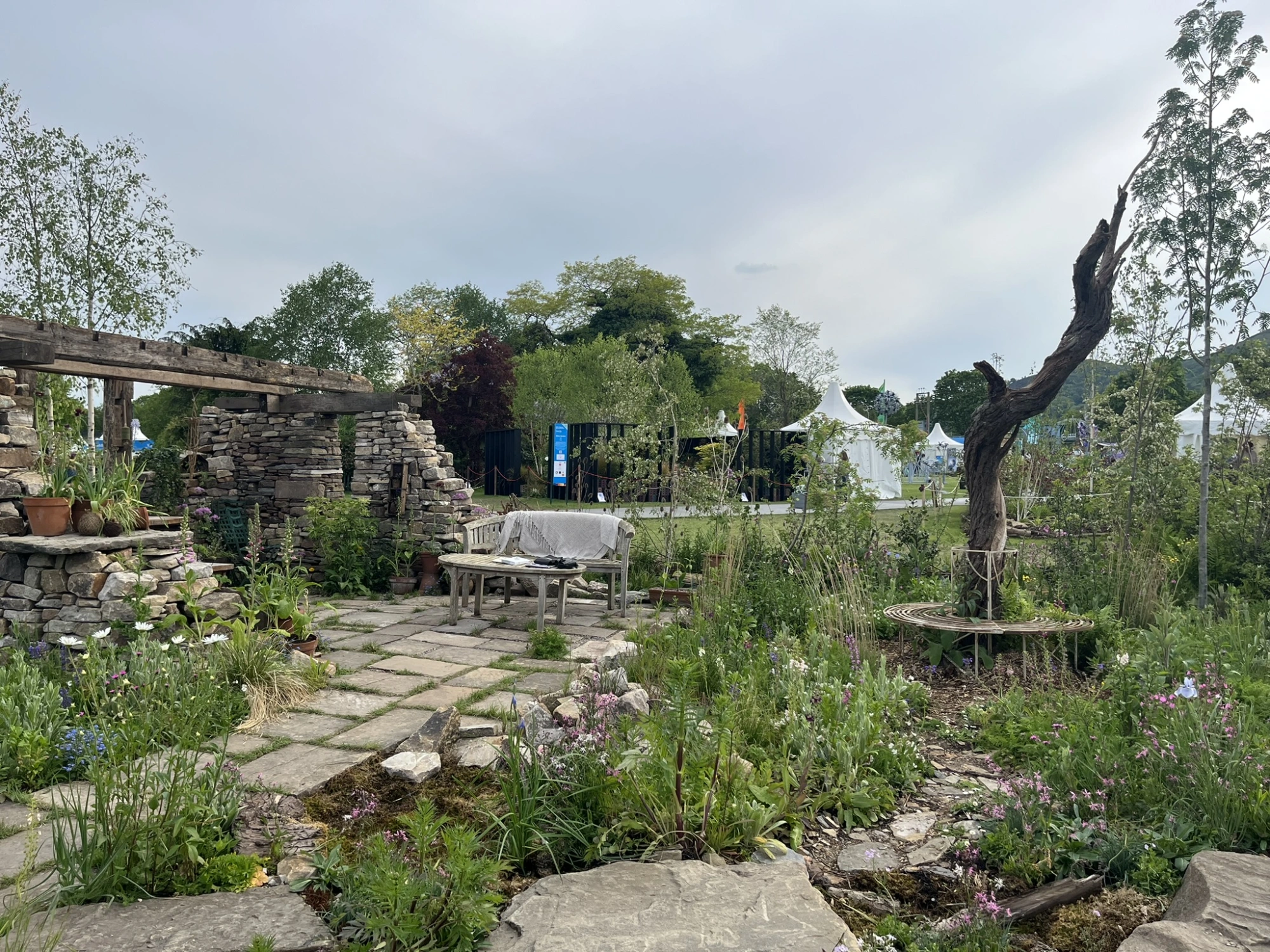A tranquil show garden featuring a rustic dry stone wall structure with wooden beams, a flagstone path, wooden seating, and abundant naturalistic planting with a twisted dead tree and a circular metal tree bench.