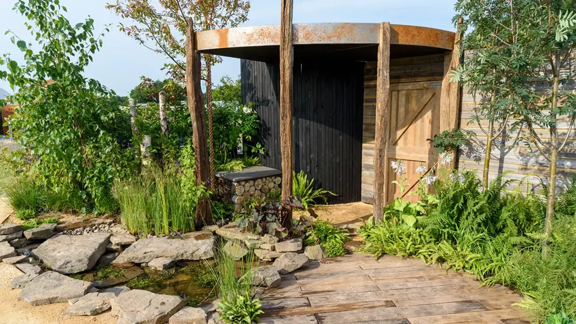 A garden features a black timber building under a rusted steel canopy, a pond with stone stepping, a wooden path, and lush ferns.