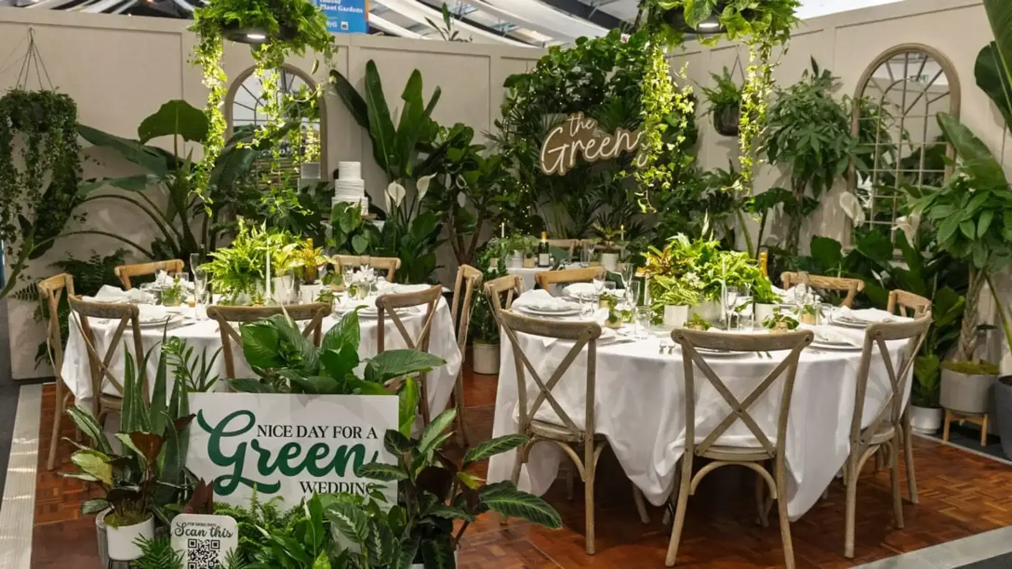 A lush indoor plant garden transformed into an elegant wedding reception setting with white dining tables, wooden cross-back chairs, and abundant green houseplants.