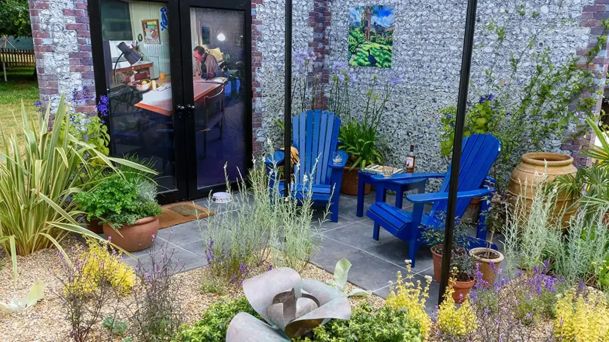 A contemporary outdoor room features a paved patio with two bright blue Adirondack chairs, surrounded by gravel paths, diverse planting, and a gabion wall.