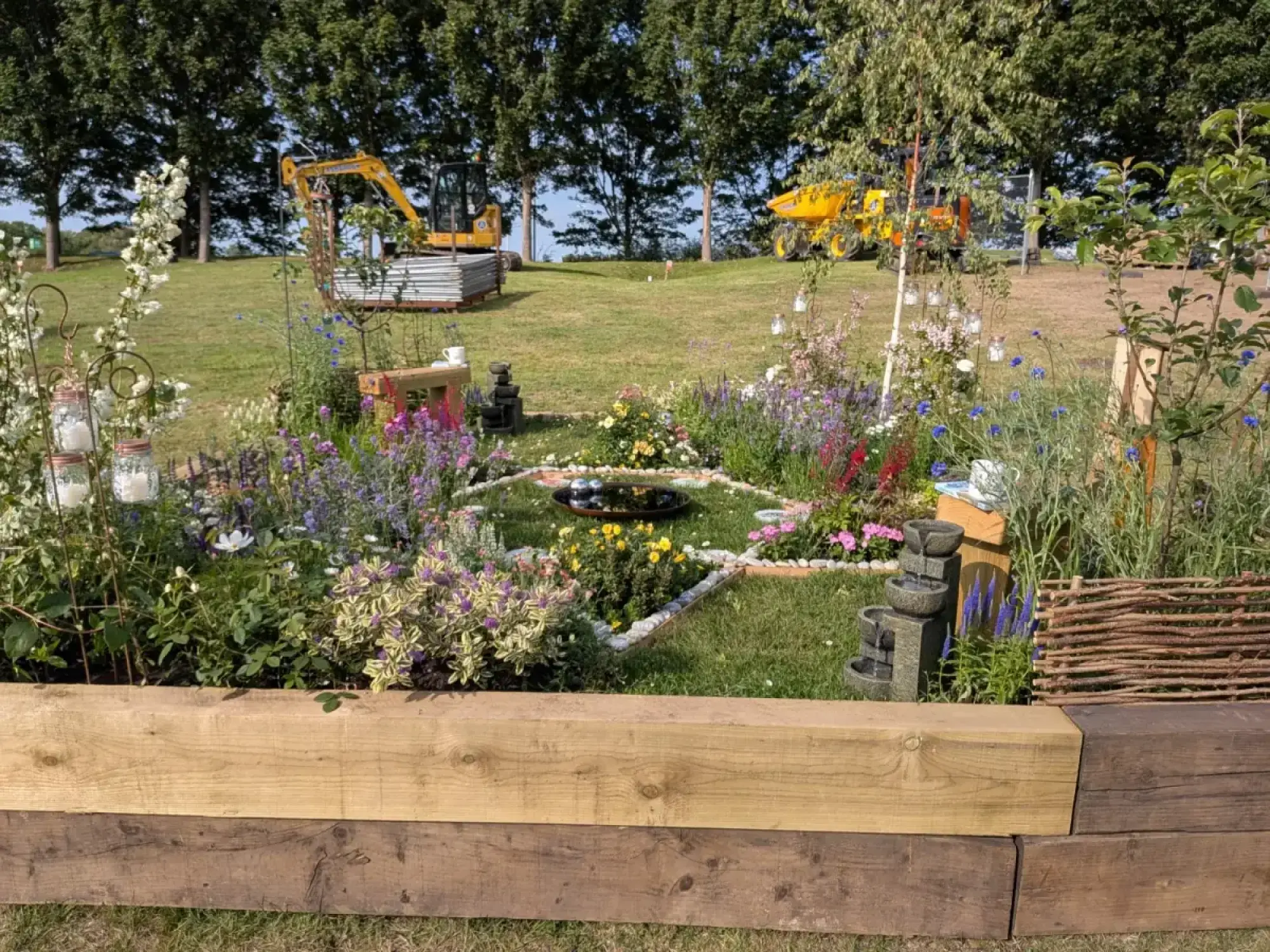 A colourful garden border featuring a central reflective pool, a tiered stone water feature, and mixed perennials, enclosed by a raised timber edge.