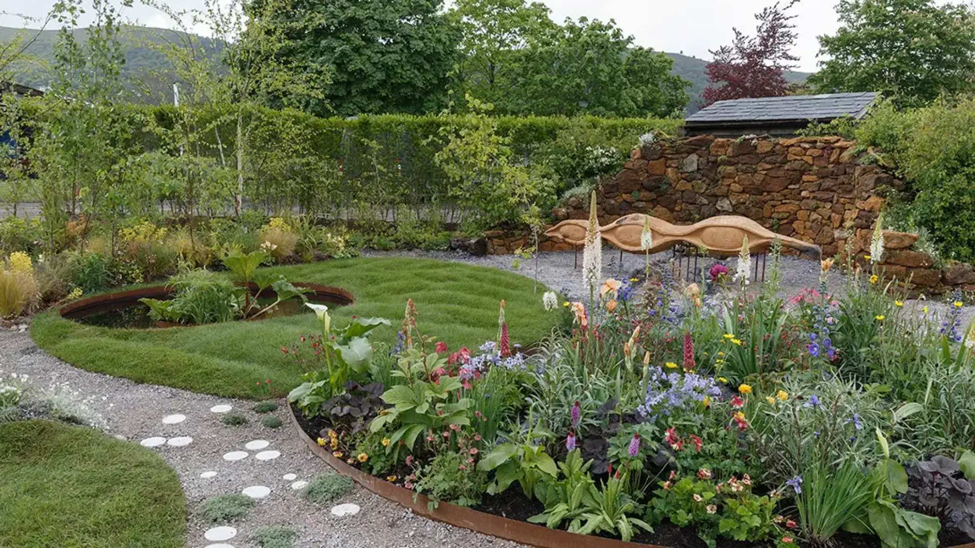 A naturalistic show garden with a carved wooden sculpture by a dry stone wall, a corten steel pond, colorful mixed perennial borders, and a gravel path.