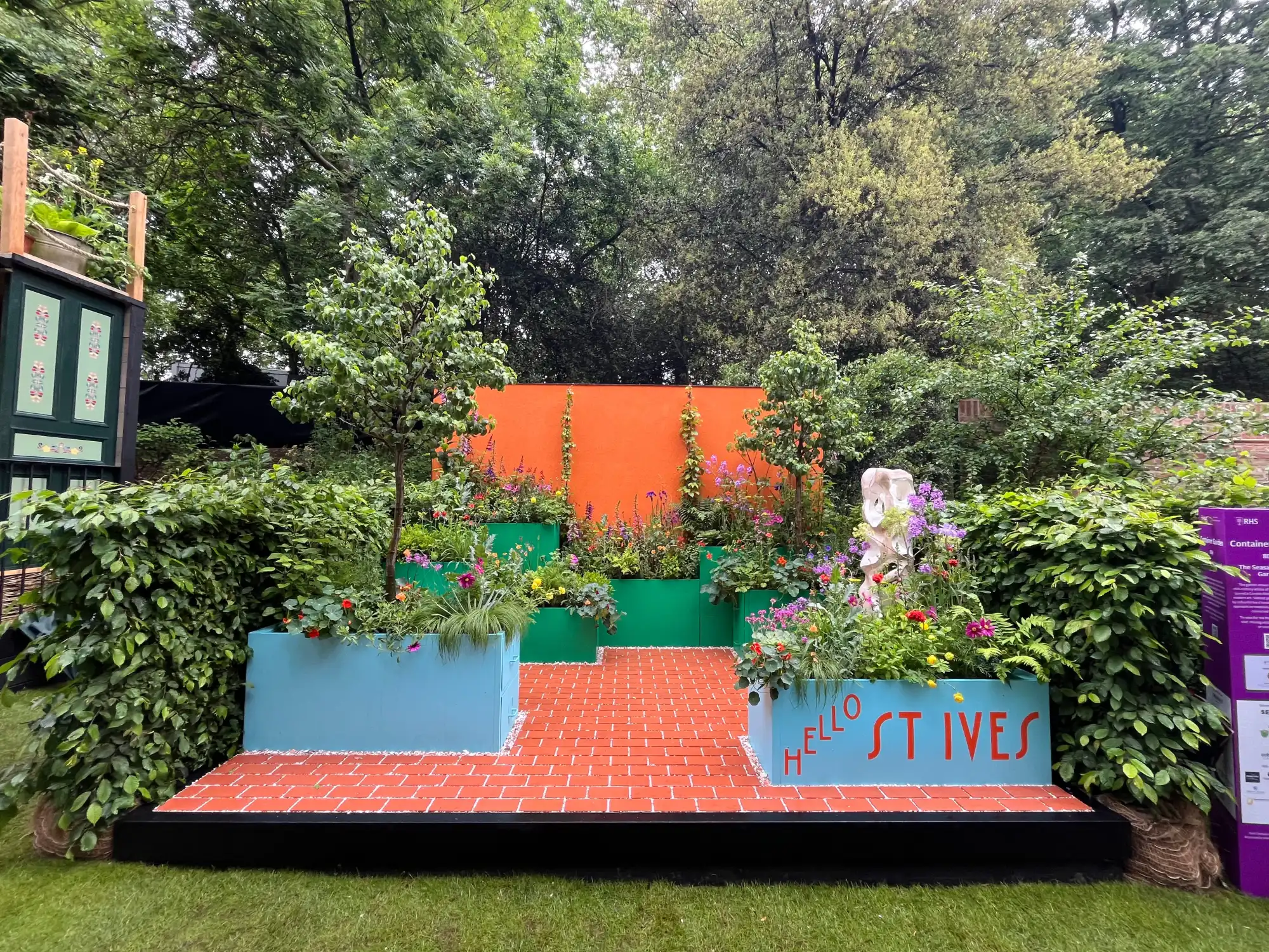 A vibrant container garden with teal raised planters spelling 'HELLO ST IVES', an orange wall, terracotta-effect paving, and colourful mixed flowers.