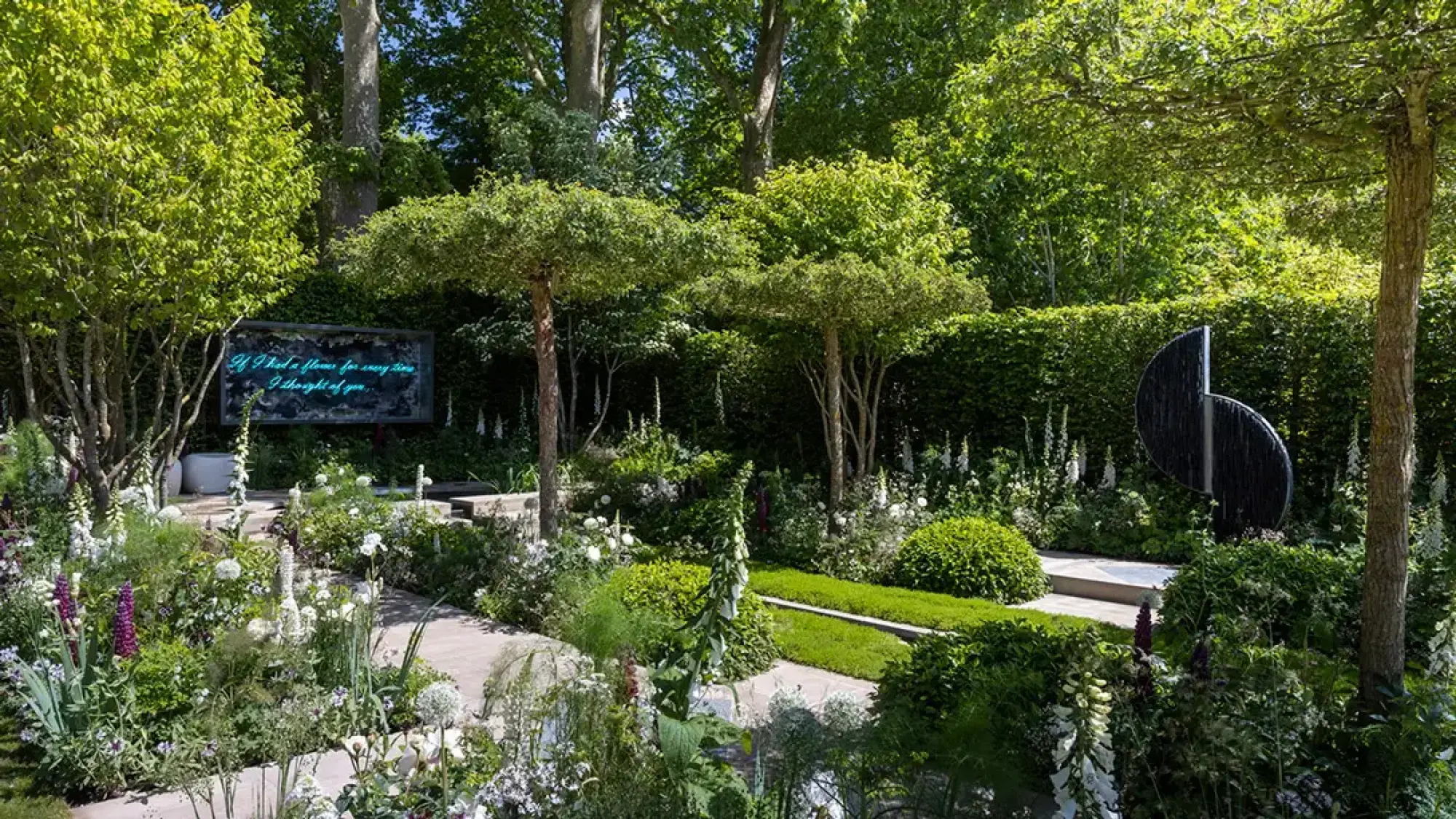 A Perennial Show Garden features a glowing screen, black abstract sculpture, umbrella trees, and paved paths through lush white and purple flowers.